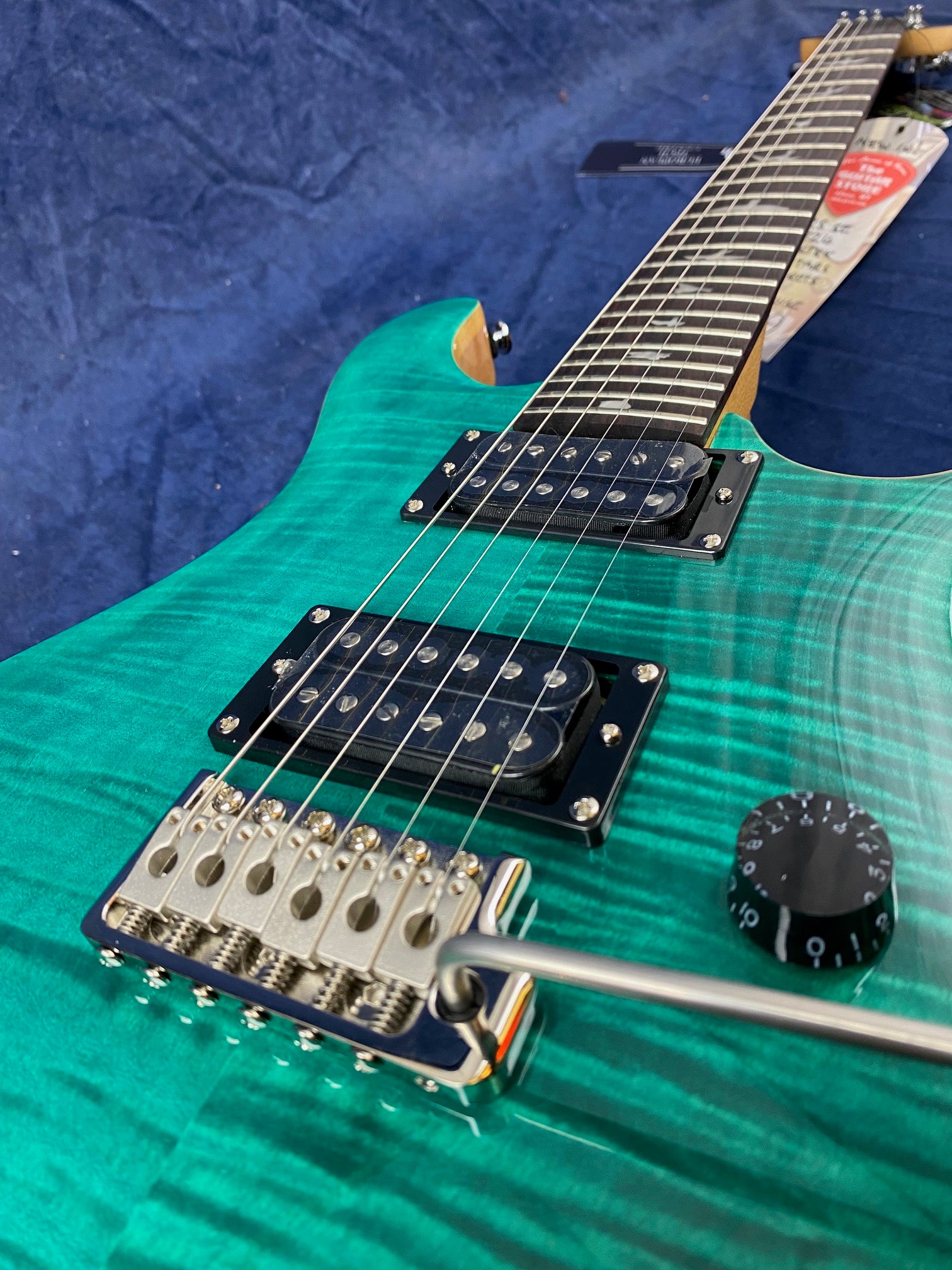 A teal electric guitar with a flamed finish, two humbucker pickups, volume knob, and a chrome tremolo bridge with whammy bar.