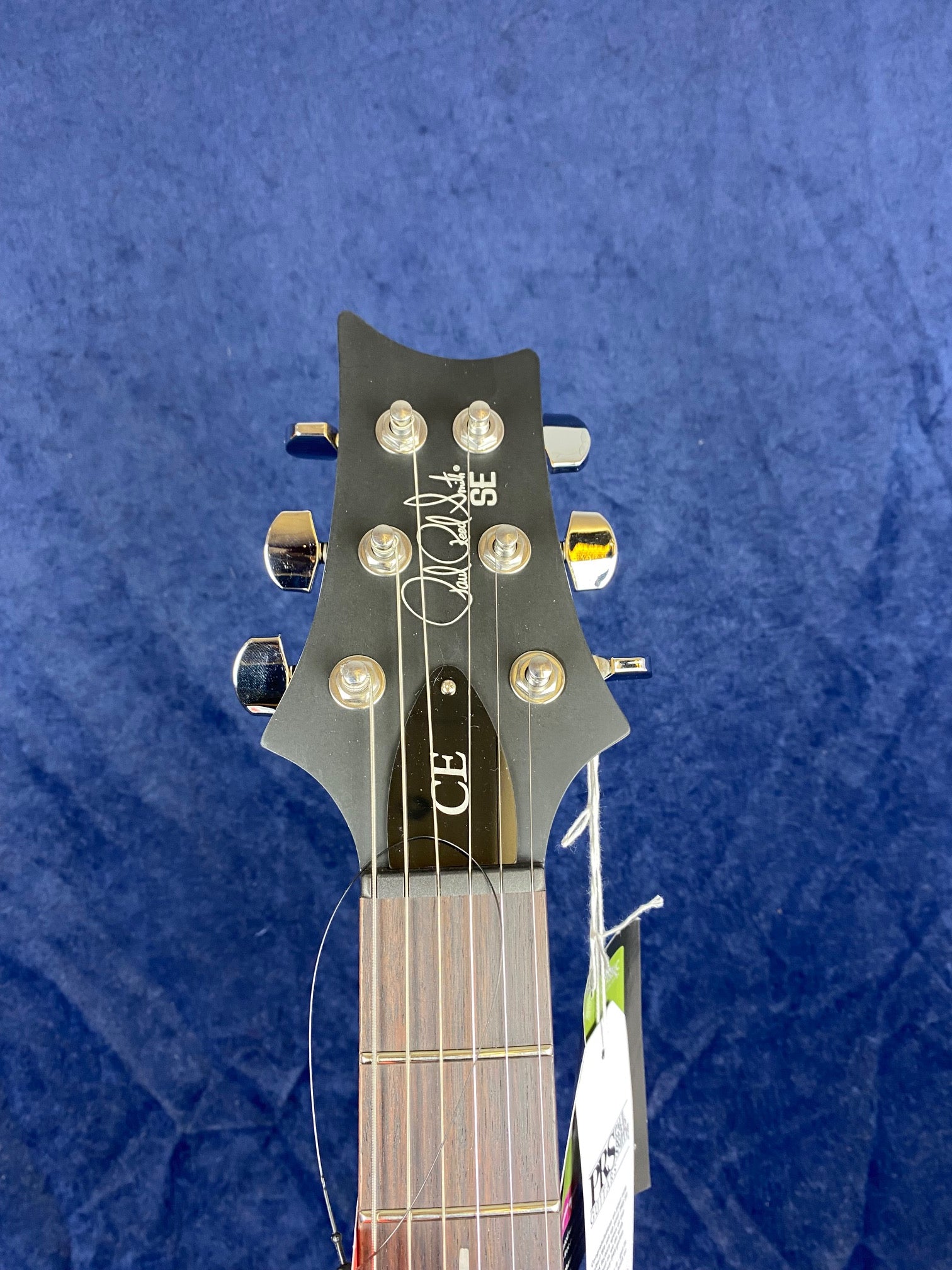 Electric guitar headstock with six tuning pegs, dark wood finish, silver hardware, and tags attached to the neck against a blue background.