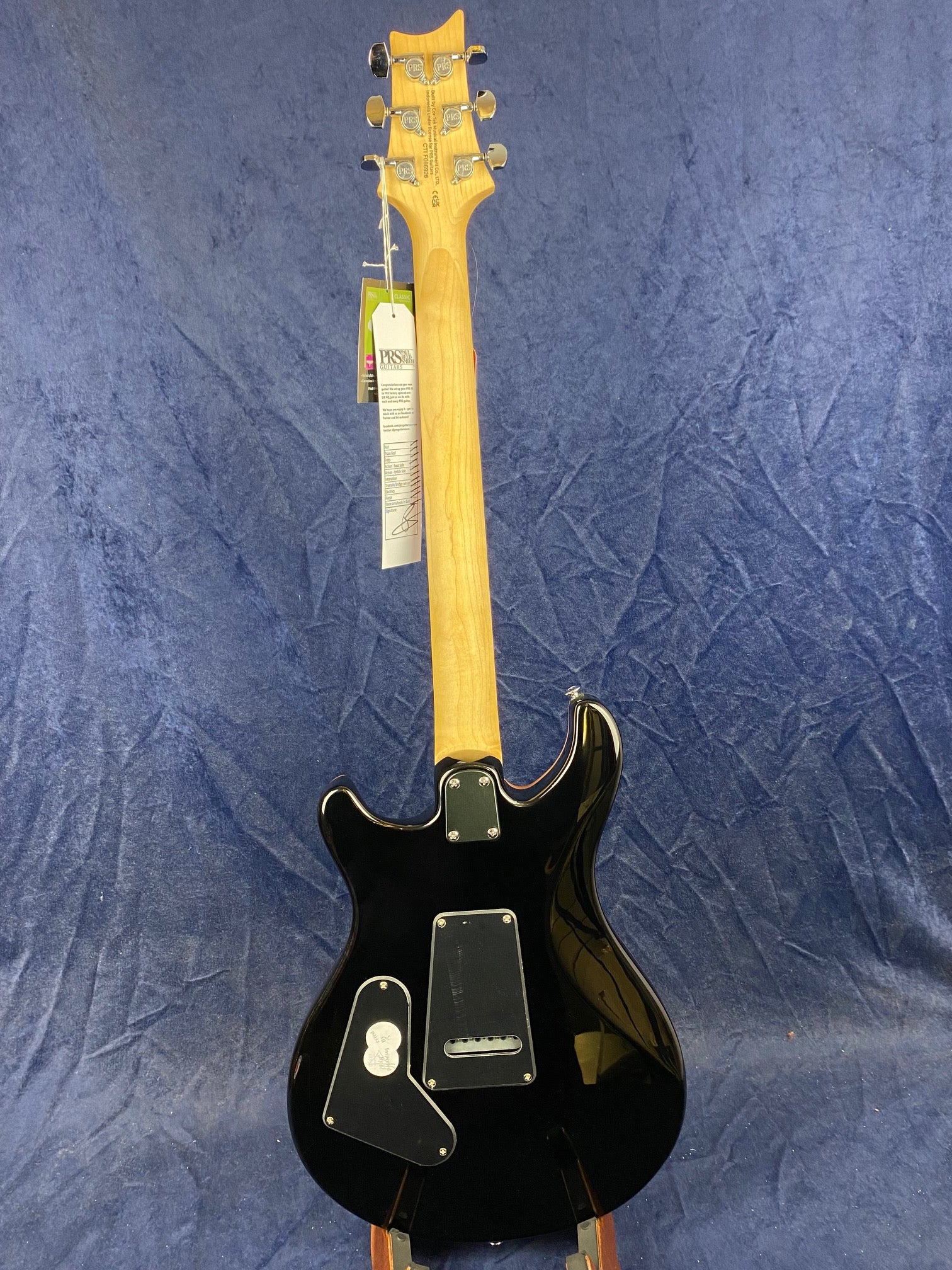 Electric guitar shown from the back, featuring a natural wood neck, black glossy body, and chrome tuners, with tags hanging from the headstock.