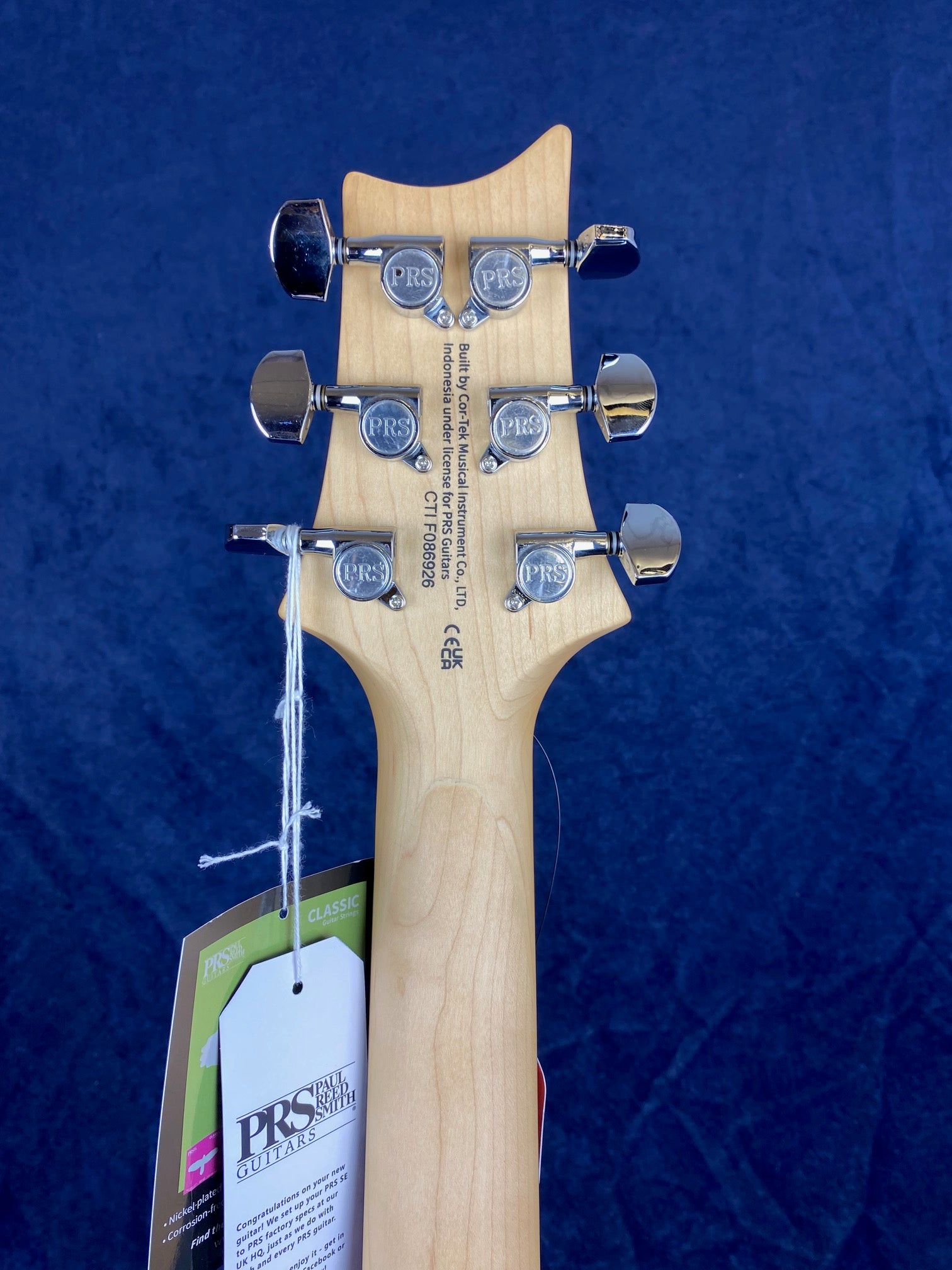 Back of a Paul Reed Smith electric guitar headstock with six chrome tuning pegs and attached tags against a navy background.
