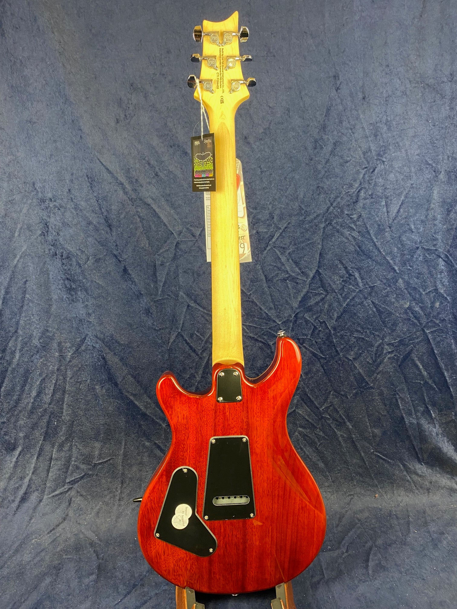 A red electric guitar shown from the back, featuring natural wood neck, black back plates, chrome tuners, and tags hanging from the headstock.