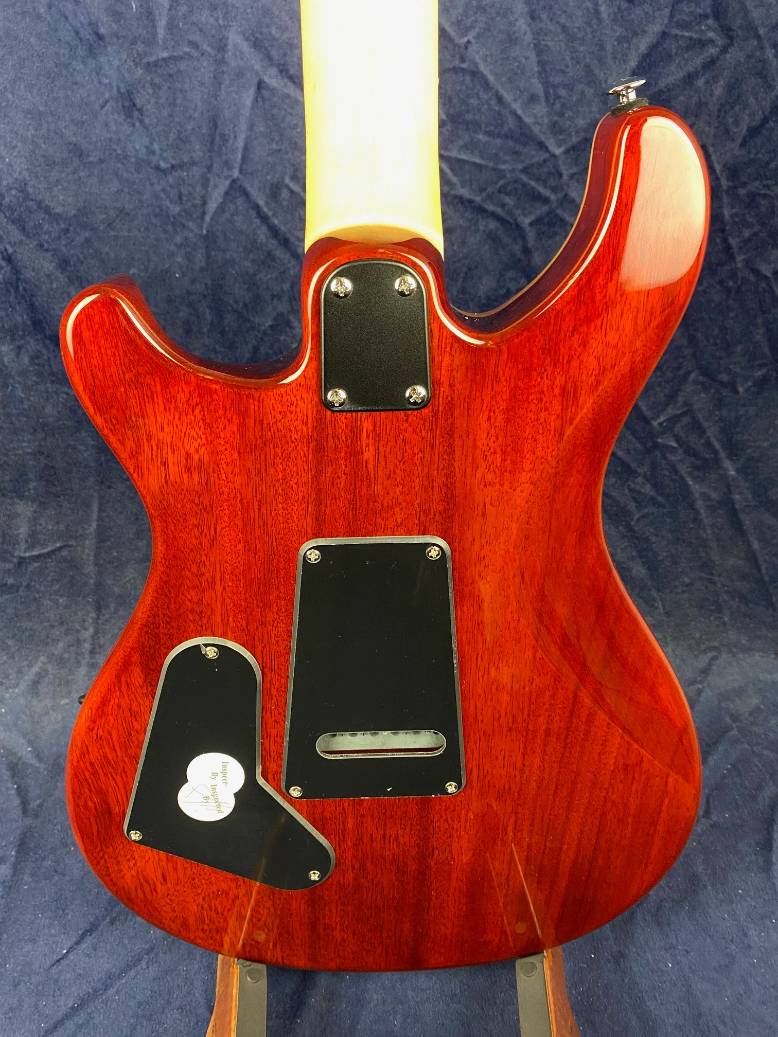 Glossy red electric guitar back with black rectangular and triangular plastic covers and a pale wood neck against a dark blue background.