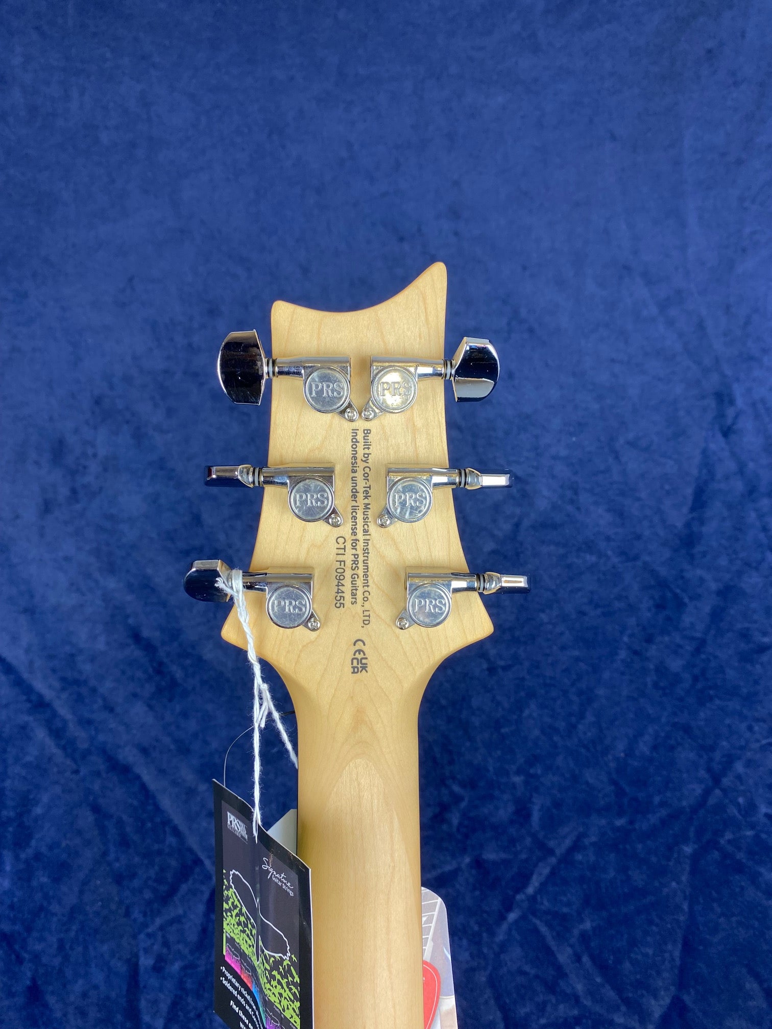 Six chrome PRS-branded tuning pegs on the back of a natural wood electric guitar headstock, with tags attached by a white string.