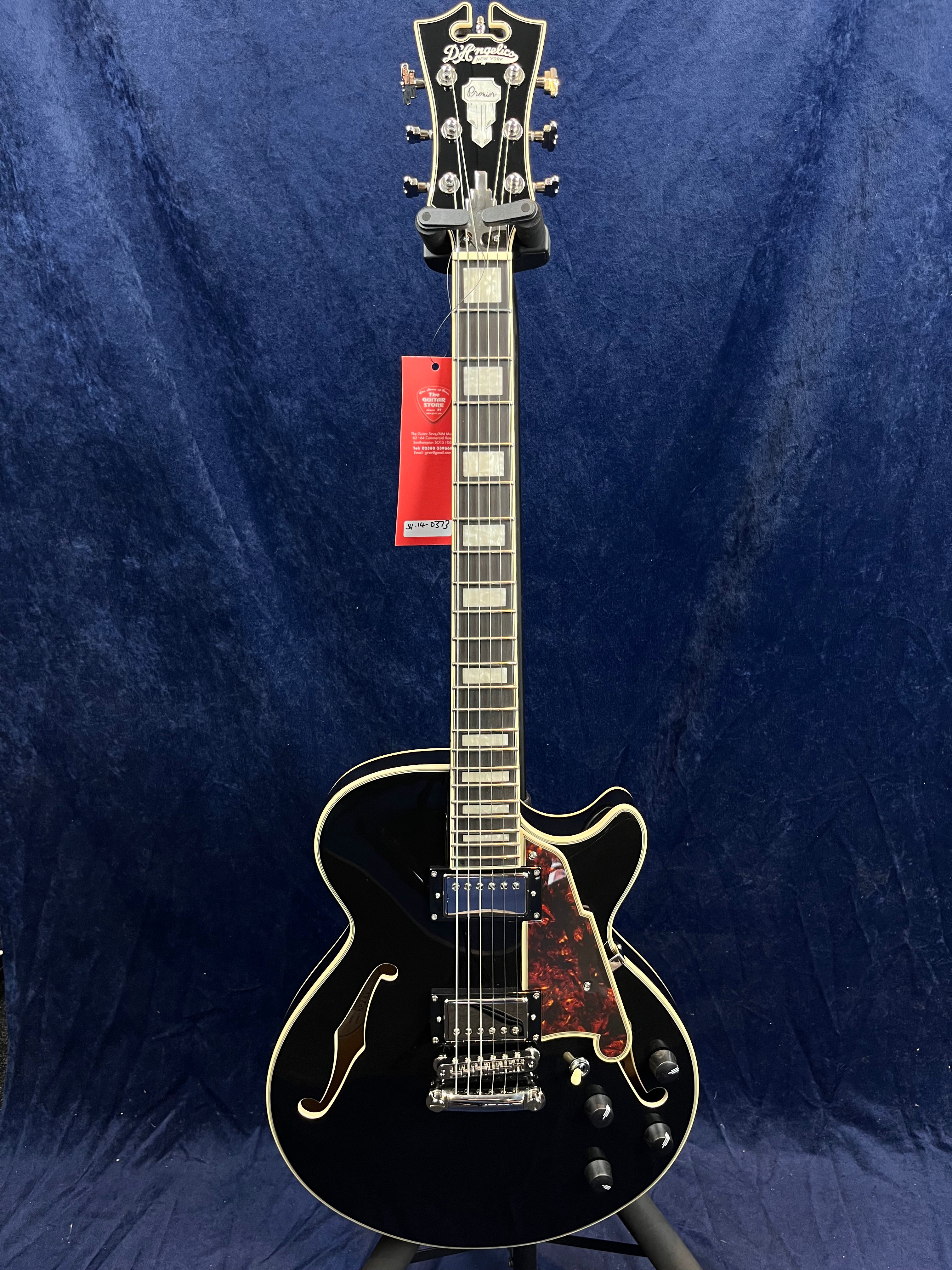 Glossy black electric guitar with cream trim, two humbucker pickups, f-holes, and a red tortoiseshell pickguard, standing against a dark blue backdrop.