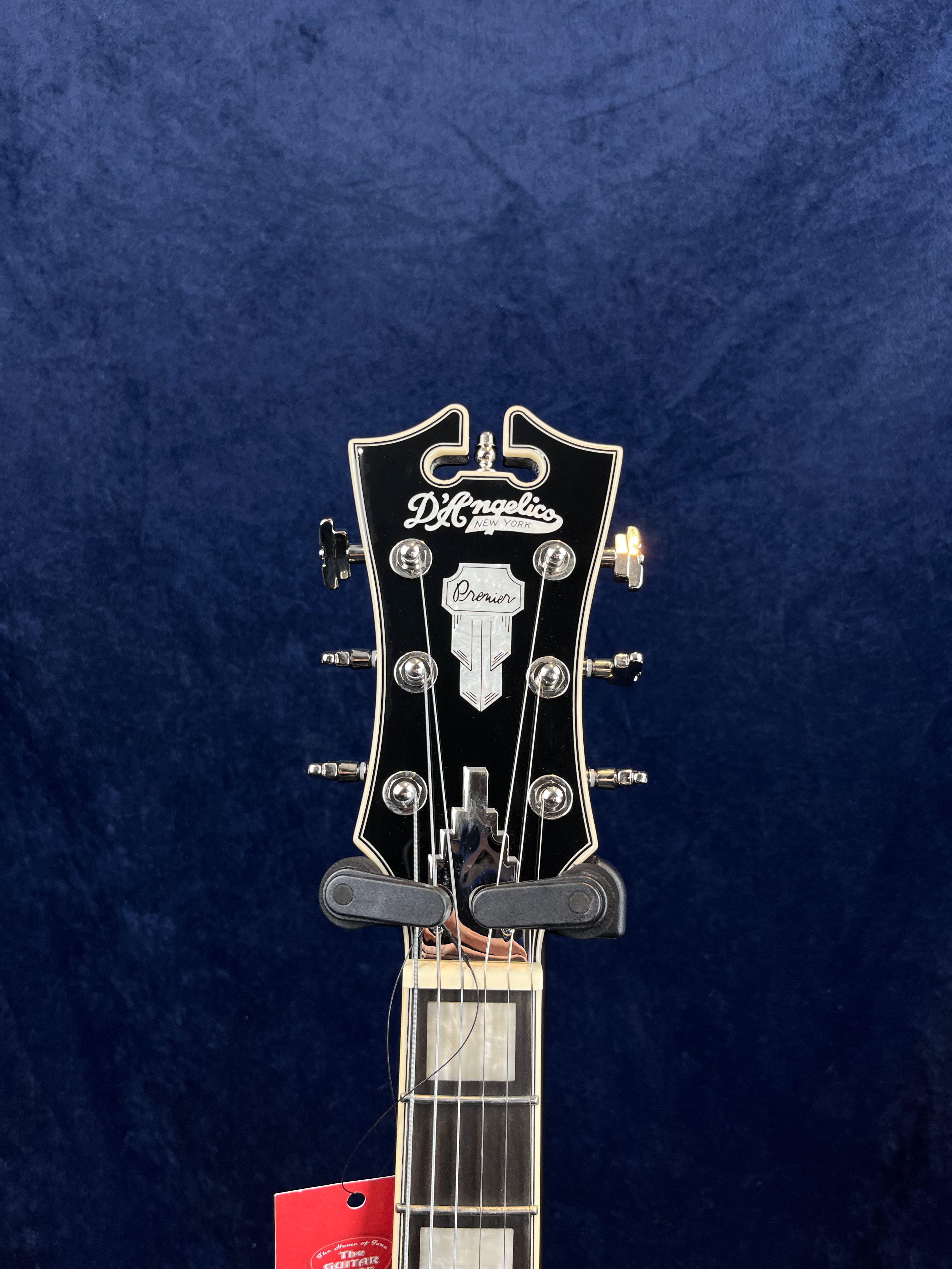 The headstock of a D’Angelico electric guitar with silver tuners and ornate inlay, a red tag hanging from the neck.