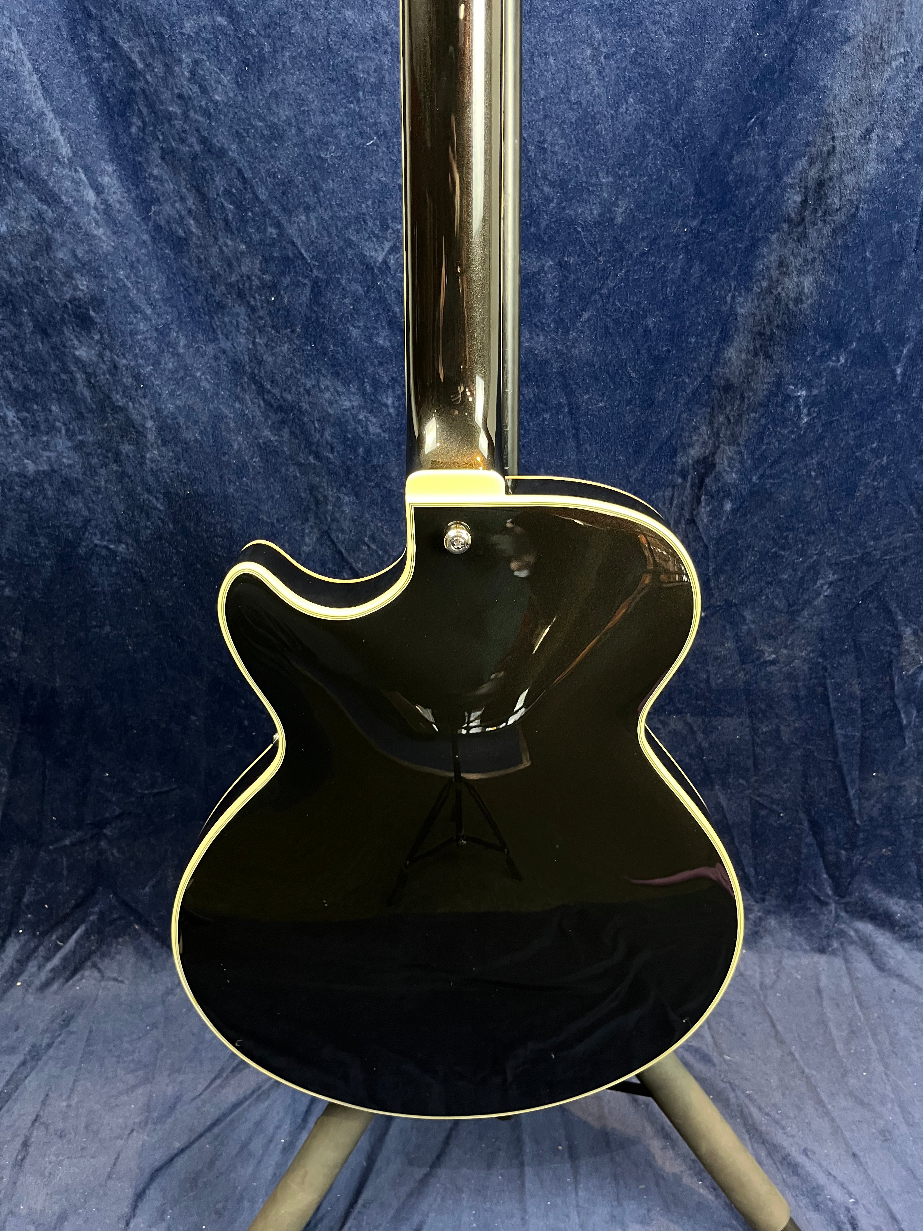 Glossy black electric guitar viewed from the back, cream binding outlining the body, single cutaway shape, standing on a tripod stand against a blue backdrop.
