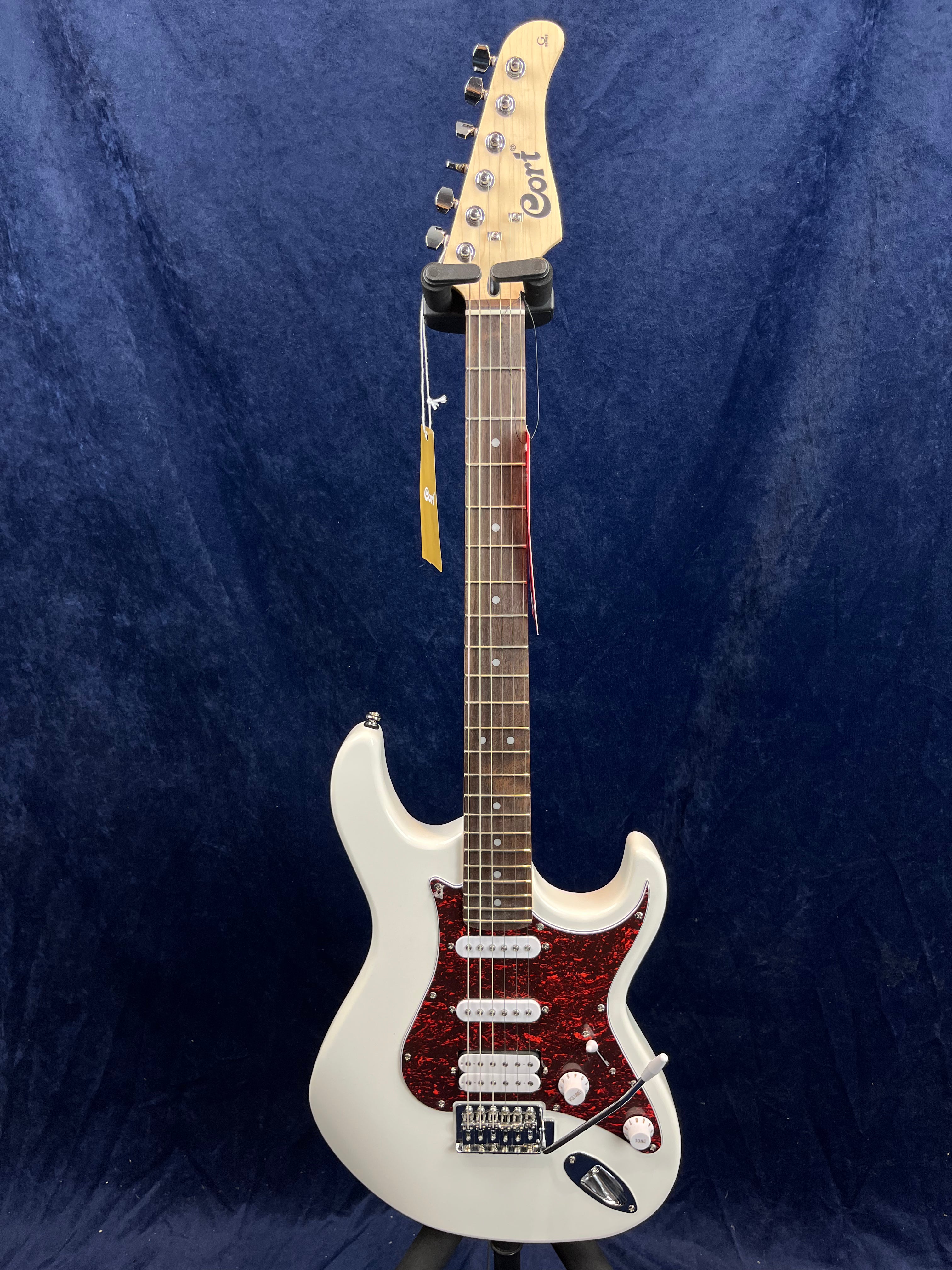 White Cort electric guitar with a dark red pickguard, three white pickups, two control knobs, and a tremolo bridge, displayed upright against a dark background.