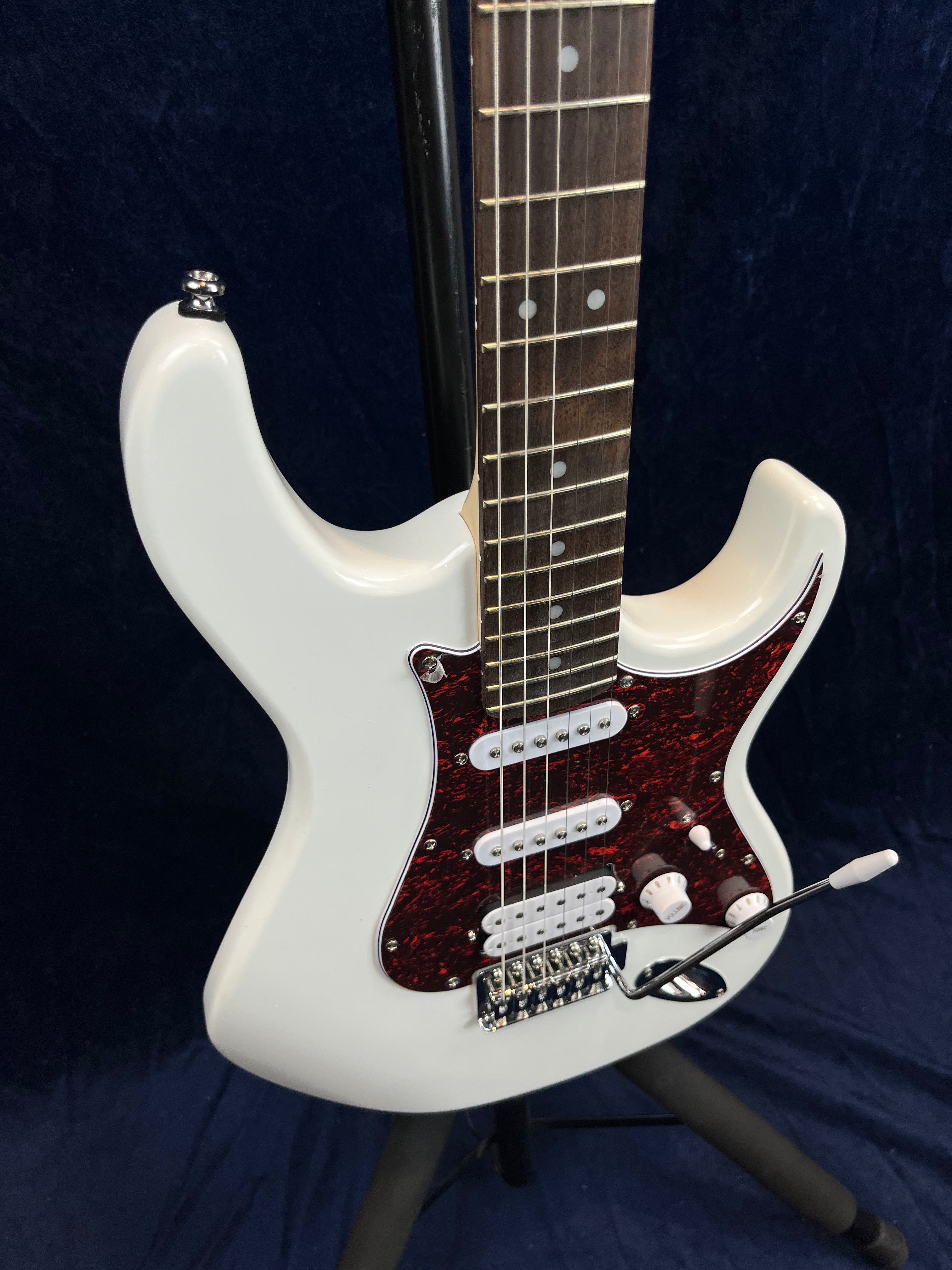 White electric guitar with a dark red pickguard, three pickups, two control knobs, and a whammy bar, resting on a stand.