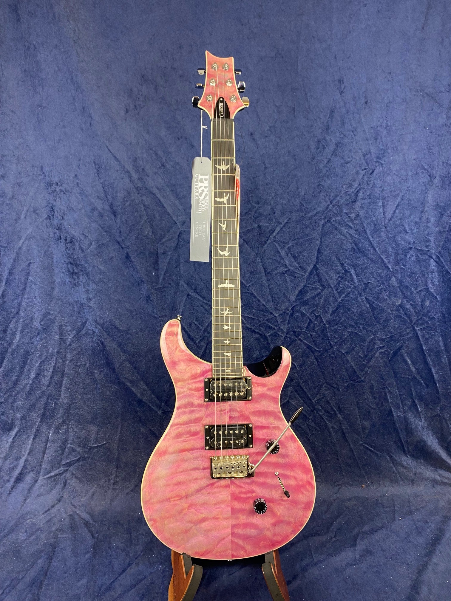 Pink electric guitar with a quilted maple top, two humbucker pickups, bird inlays on the fretboard, and a whammy bar, standing on a stand.