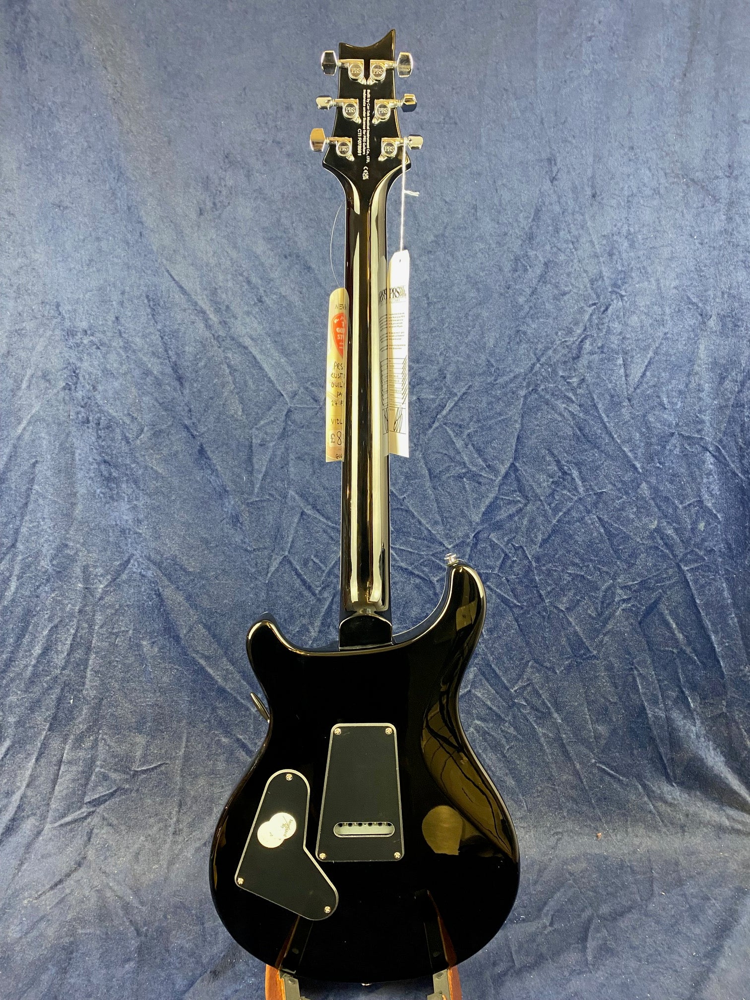 Glossy black electric guitar shown from the back, tags hanging from headstock, set against a blue textured background.