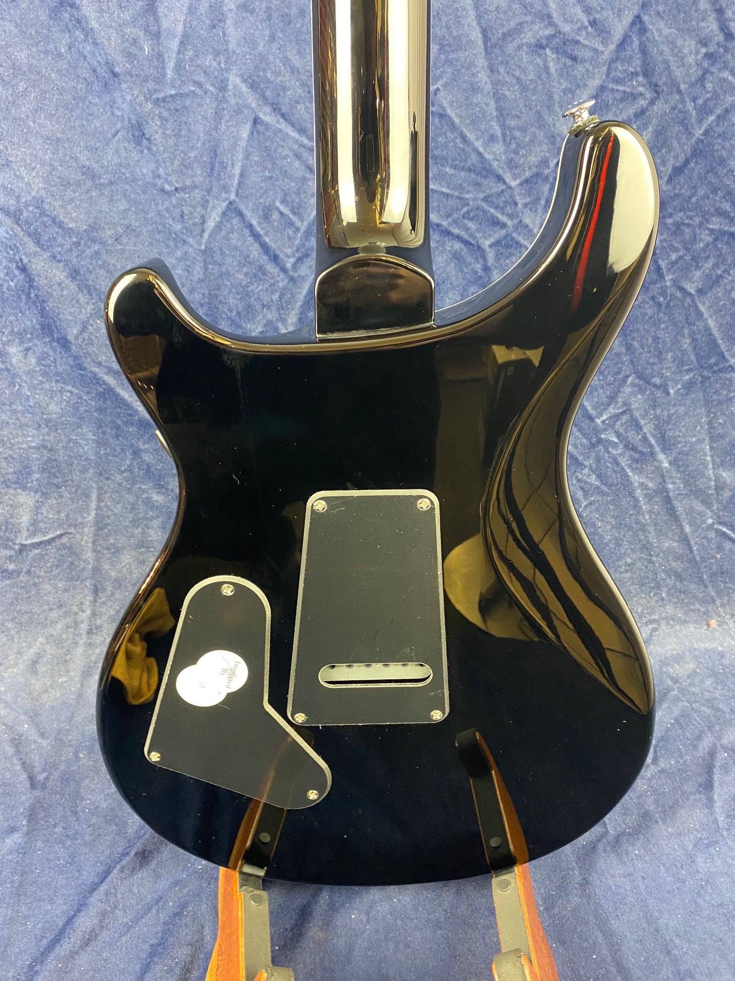 Glossy black electric guitar back, showing two access plates with screws and a neck plate, set against a blue textured background.