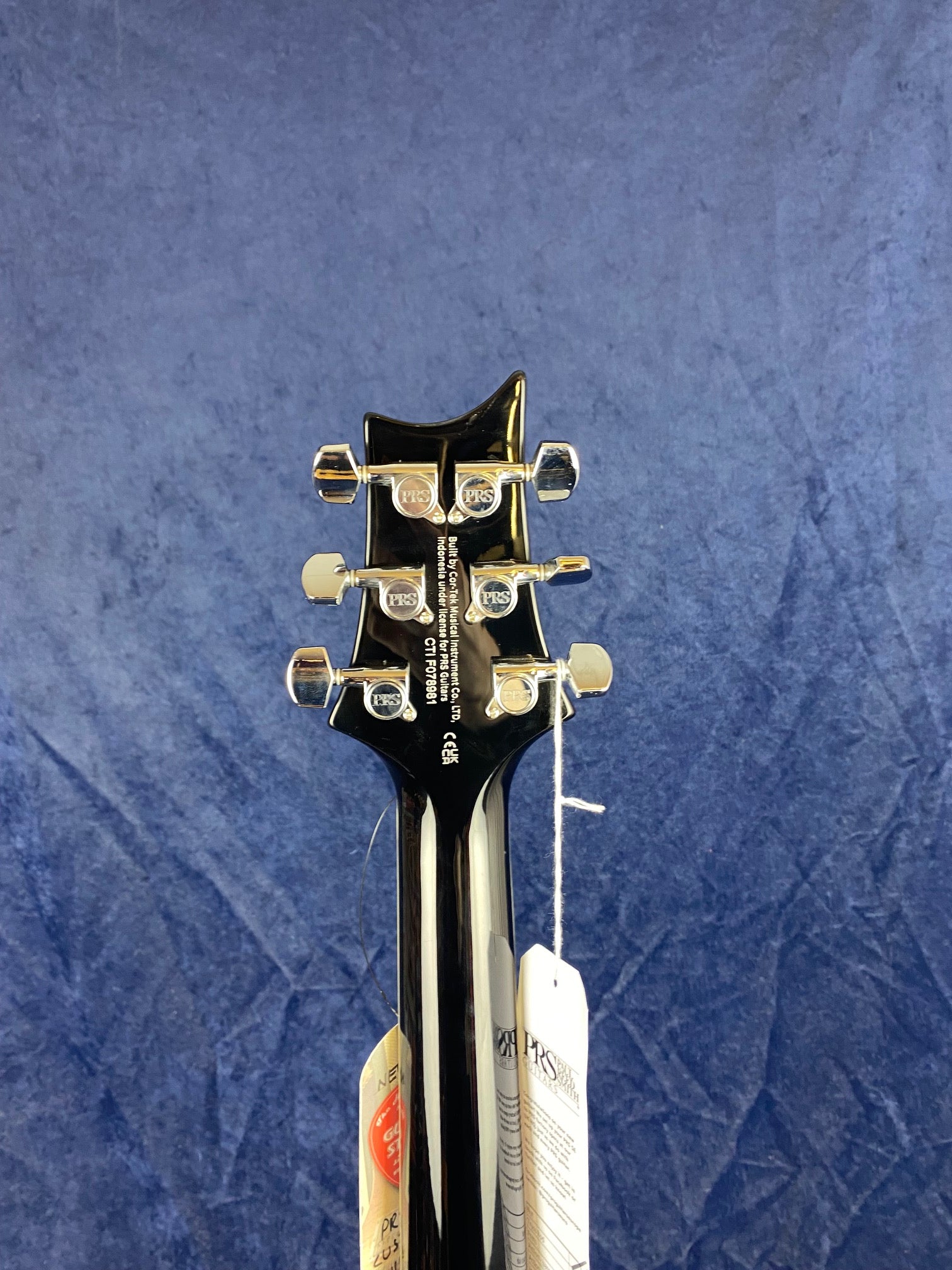 Headstock of a PRS electric guitar showing six chrome tuning pegs, black finish, and product tags hanging from the neck against a blue background.