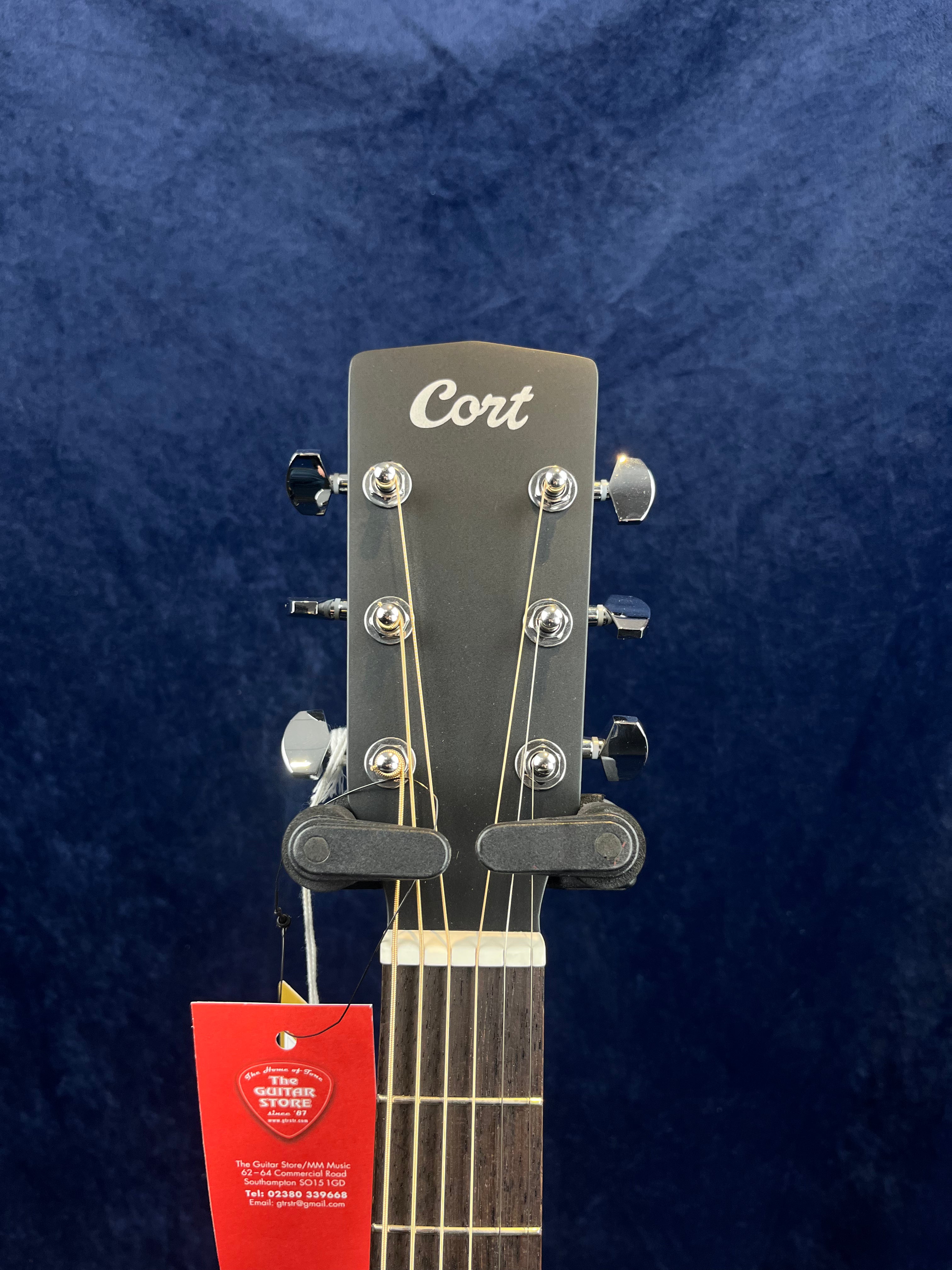 Cort acoustic guitar headstock with six tuning pegs, visible strings, and a red tag hanging from the neck against a dark blue background.