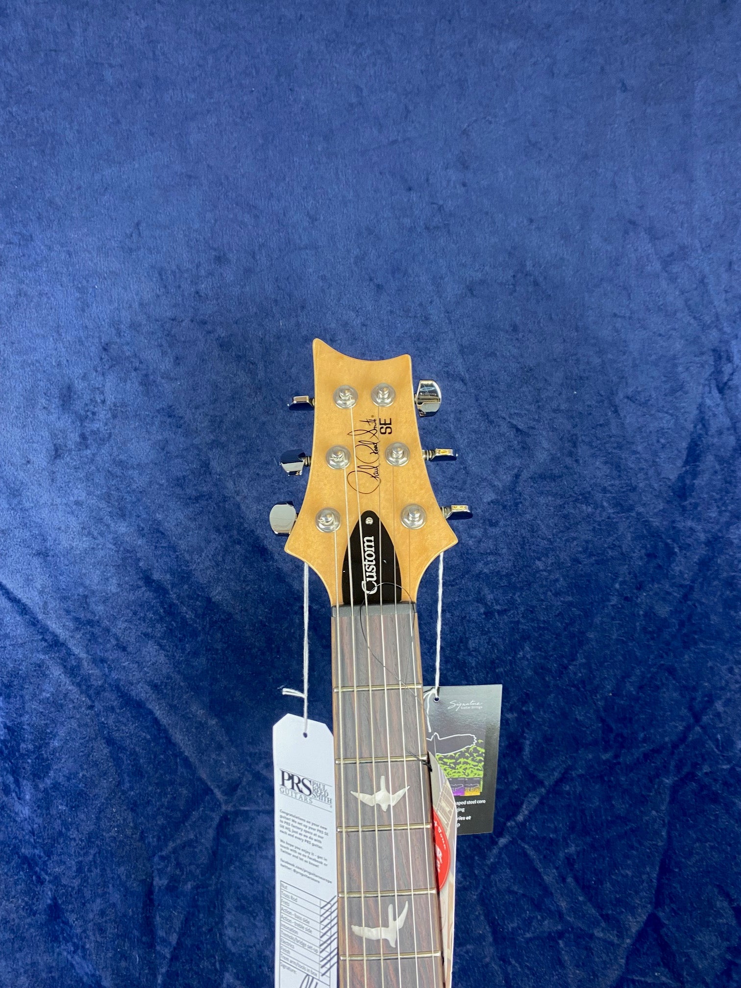 PRS SE Custom electric guitar headstock with six tuning pegs, bird inlays on the fretboard, and tags attached below the nut. Blue background.