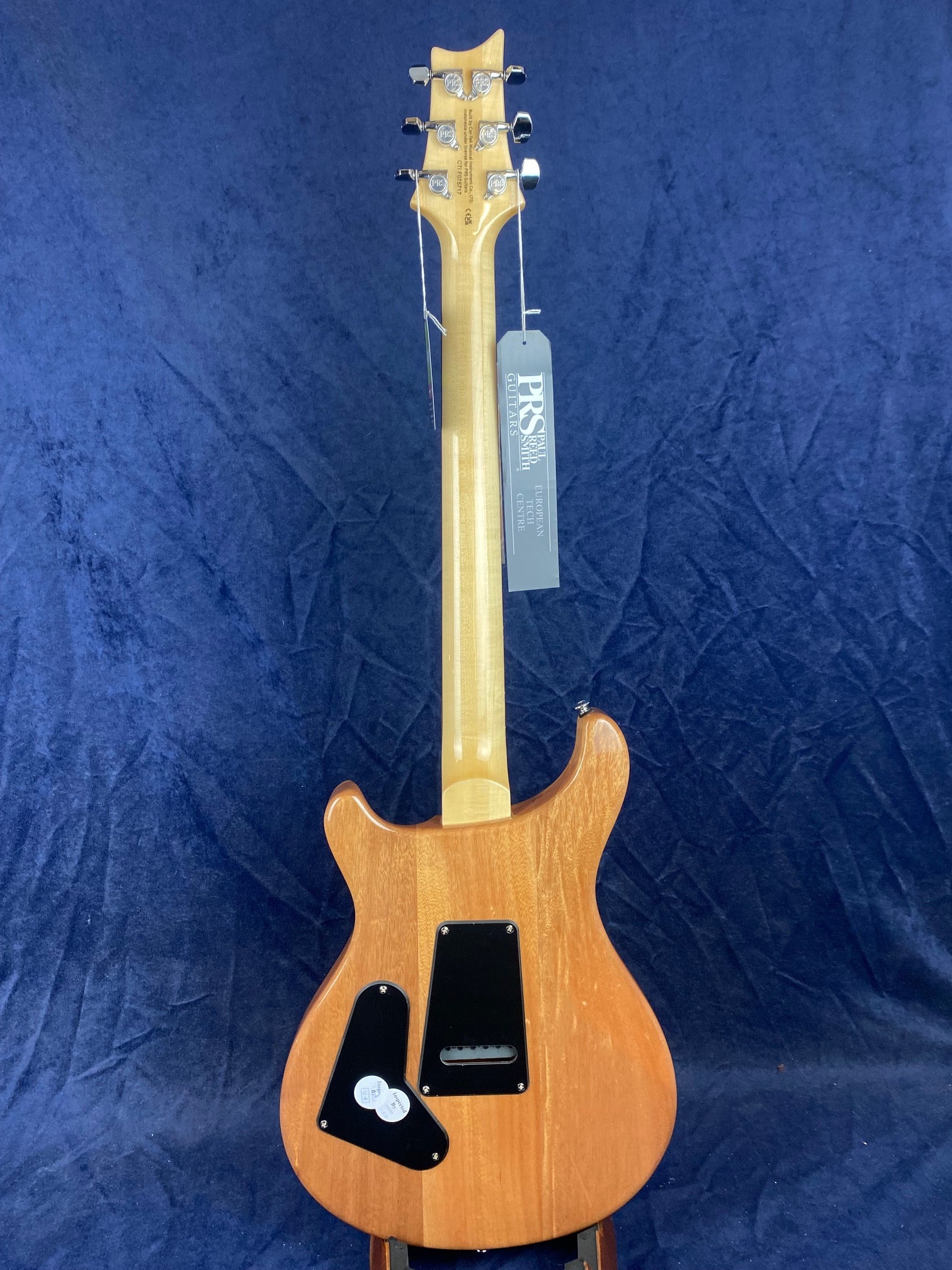 Electric guitar viewed from the back, showing a natural wood finish, two black plastic cavity covers, and a PRS Guitars tag hanging from a tuning peg.