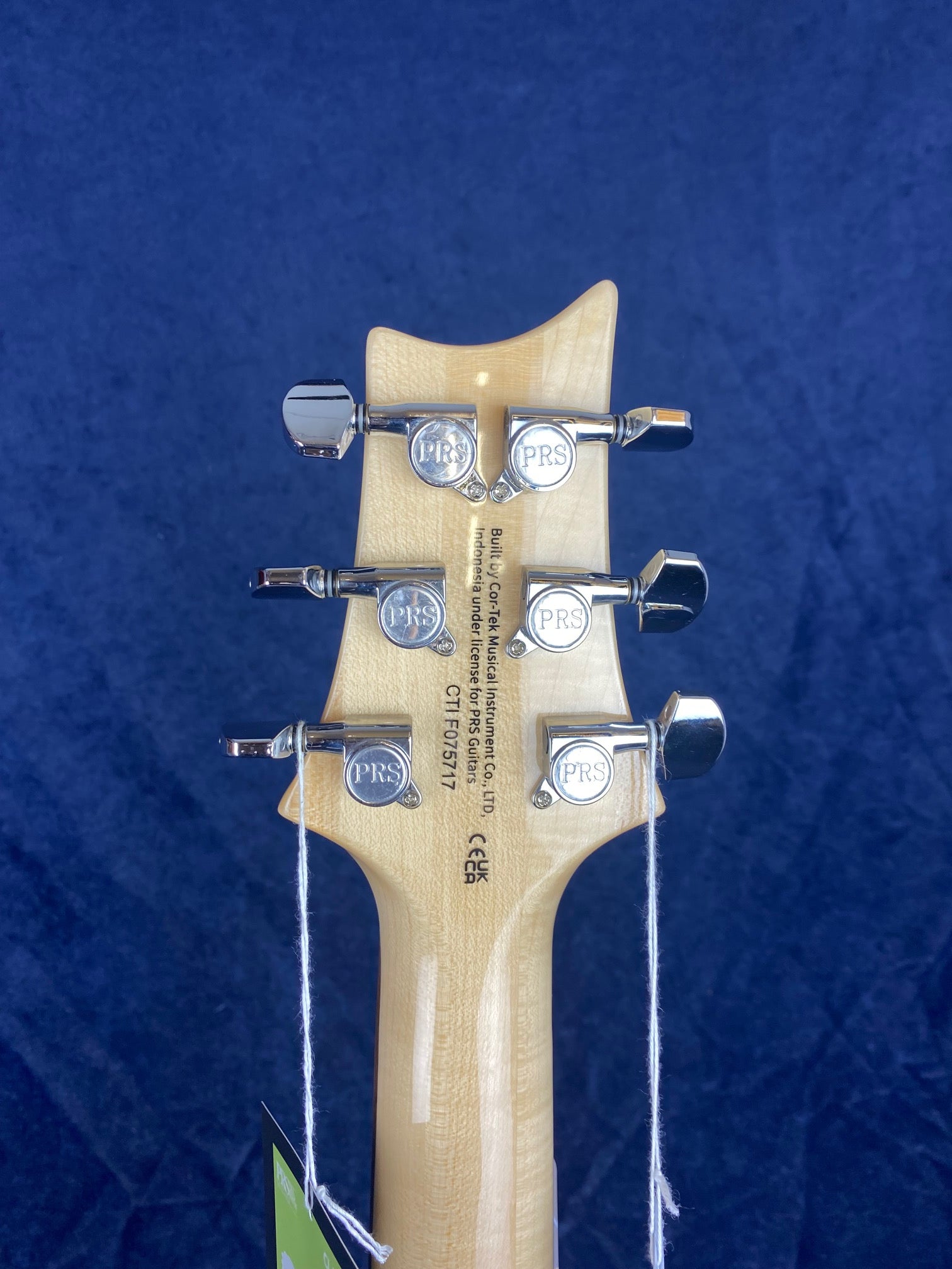 Back of a light wood electric guitar headstock with six PRS-branded chrome tuning pegs and product information printed vertically.