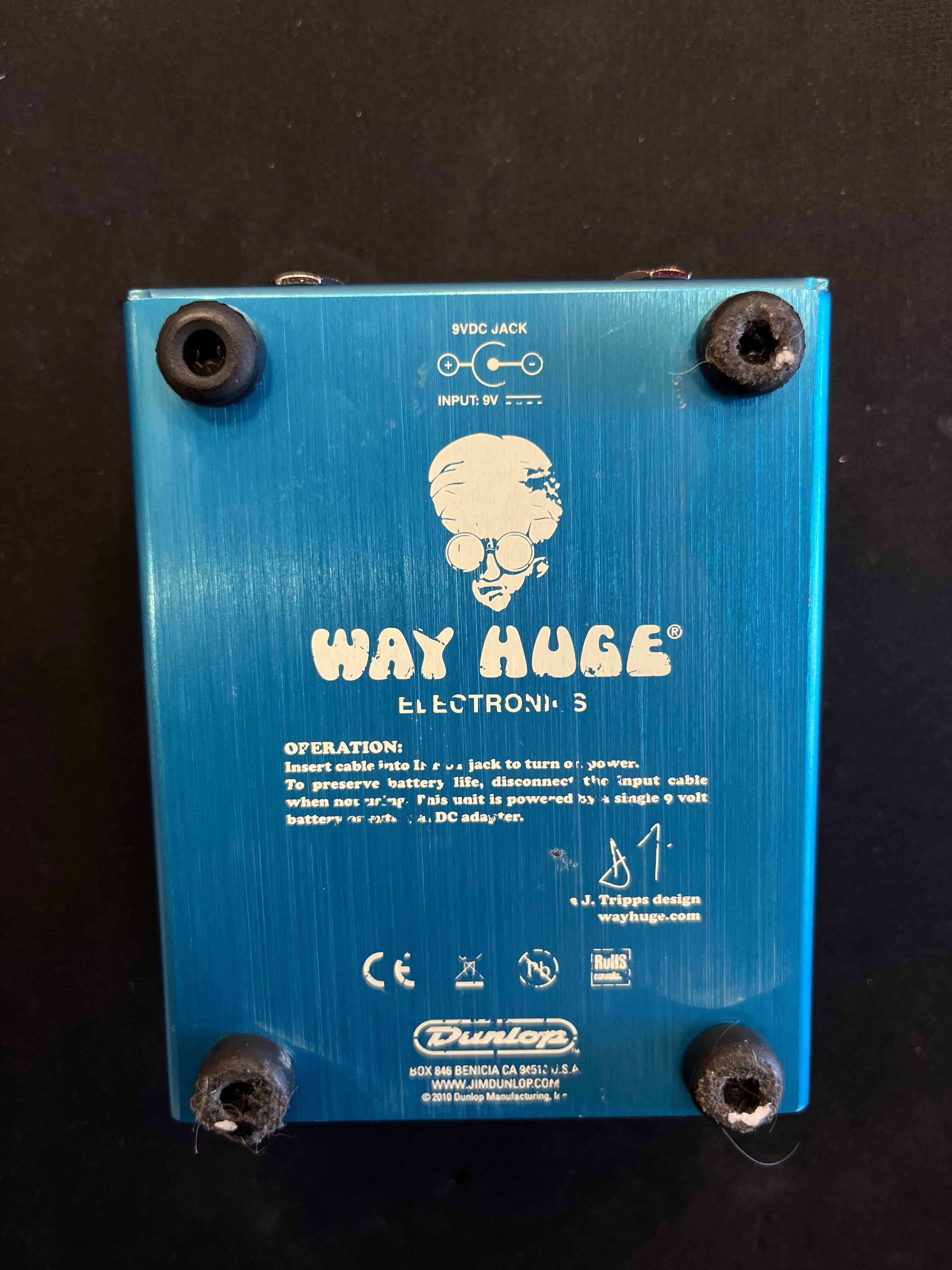 Blue metal guitar pedal bottom labeled "Way Huge Electronics," with instructions, rubber feet on corners, and a cartoon face with glasses and big hair.