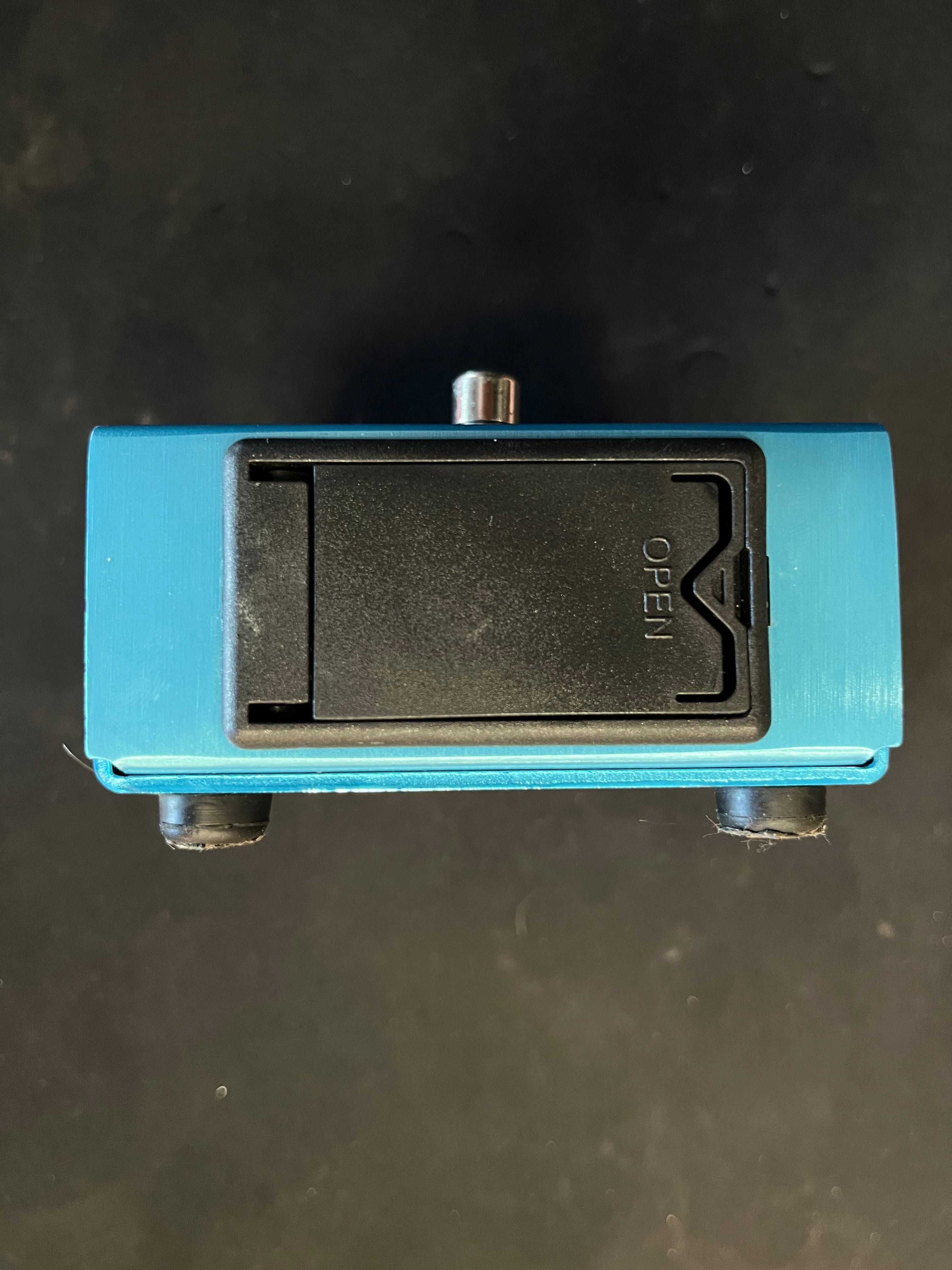 A blue pre-owned guitar effects pedal viewed from the bottom, showing a black battery compartment cover with "open" instructions.