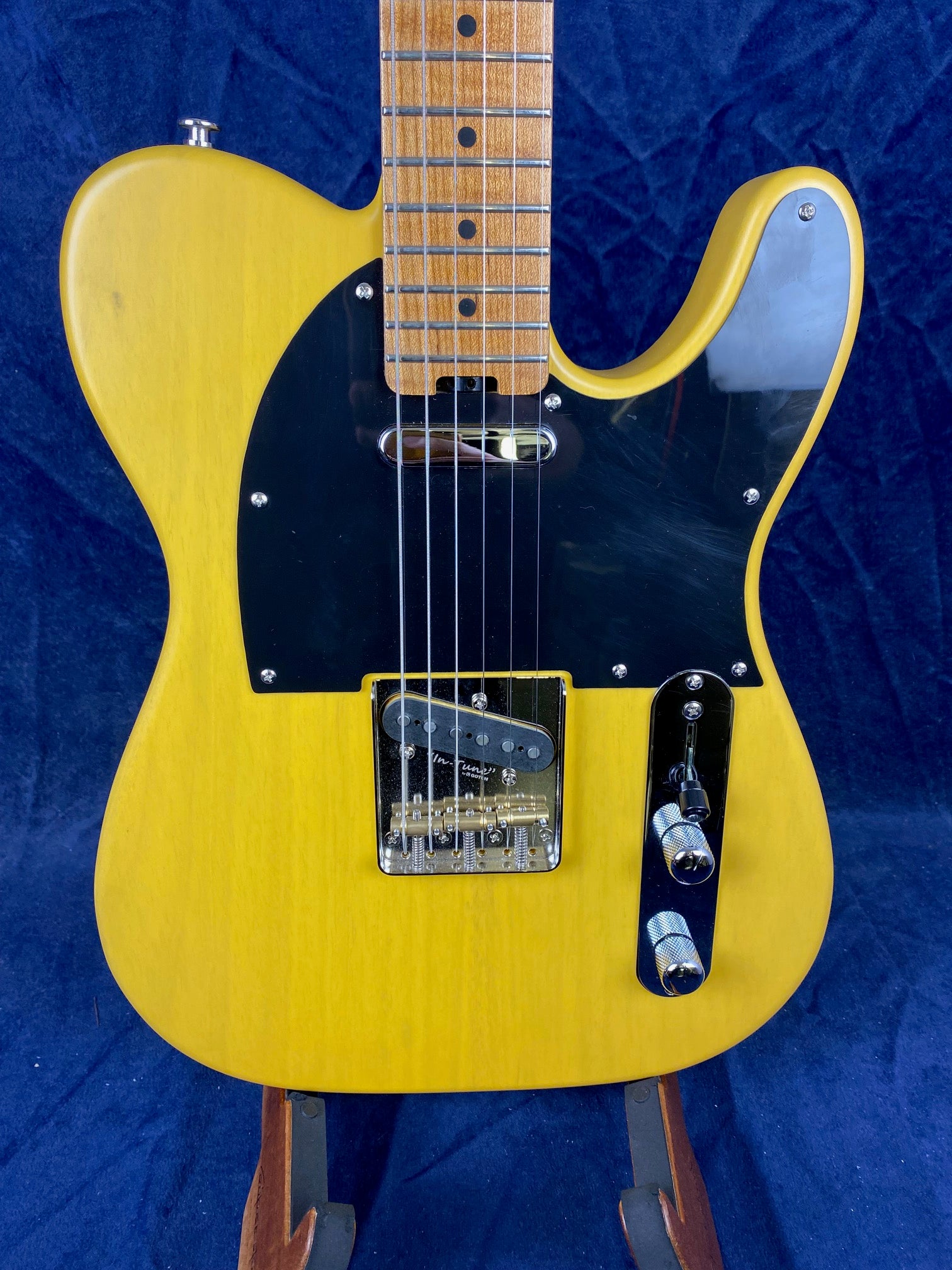 Yellow electric guitar with a black pickguard, two control knobs, single cutaway body, and a maple neck, shown on a stand against a blue background.