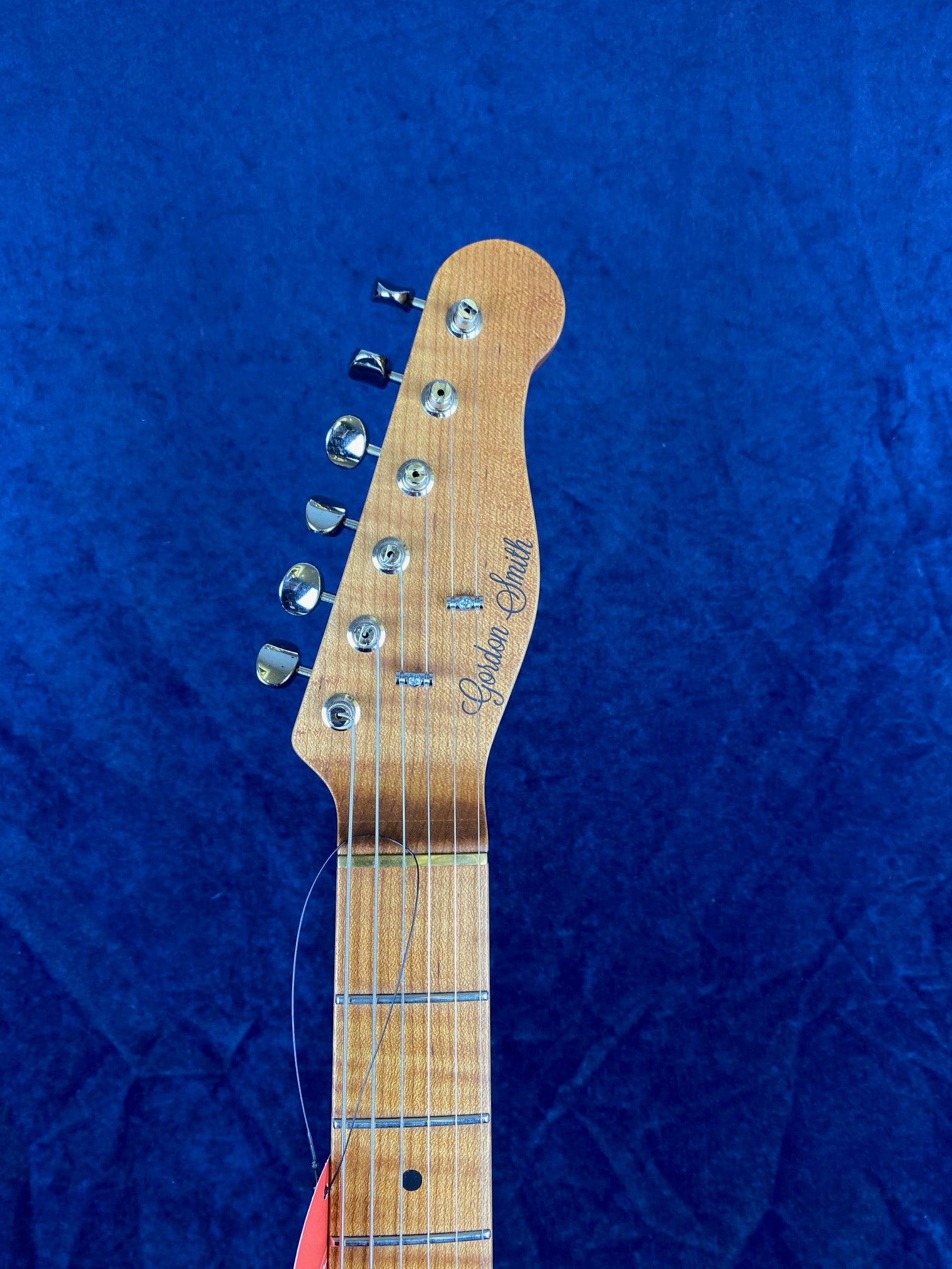 Light wood electric guitar headstock with six chrome tuning pegs, visible strings, and "Gordon Smith" written in script on the front. Blue background.