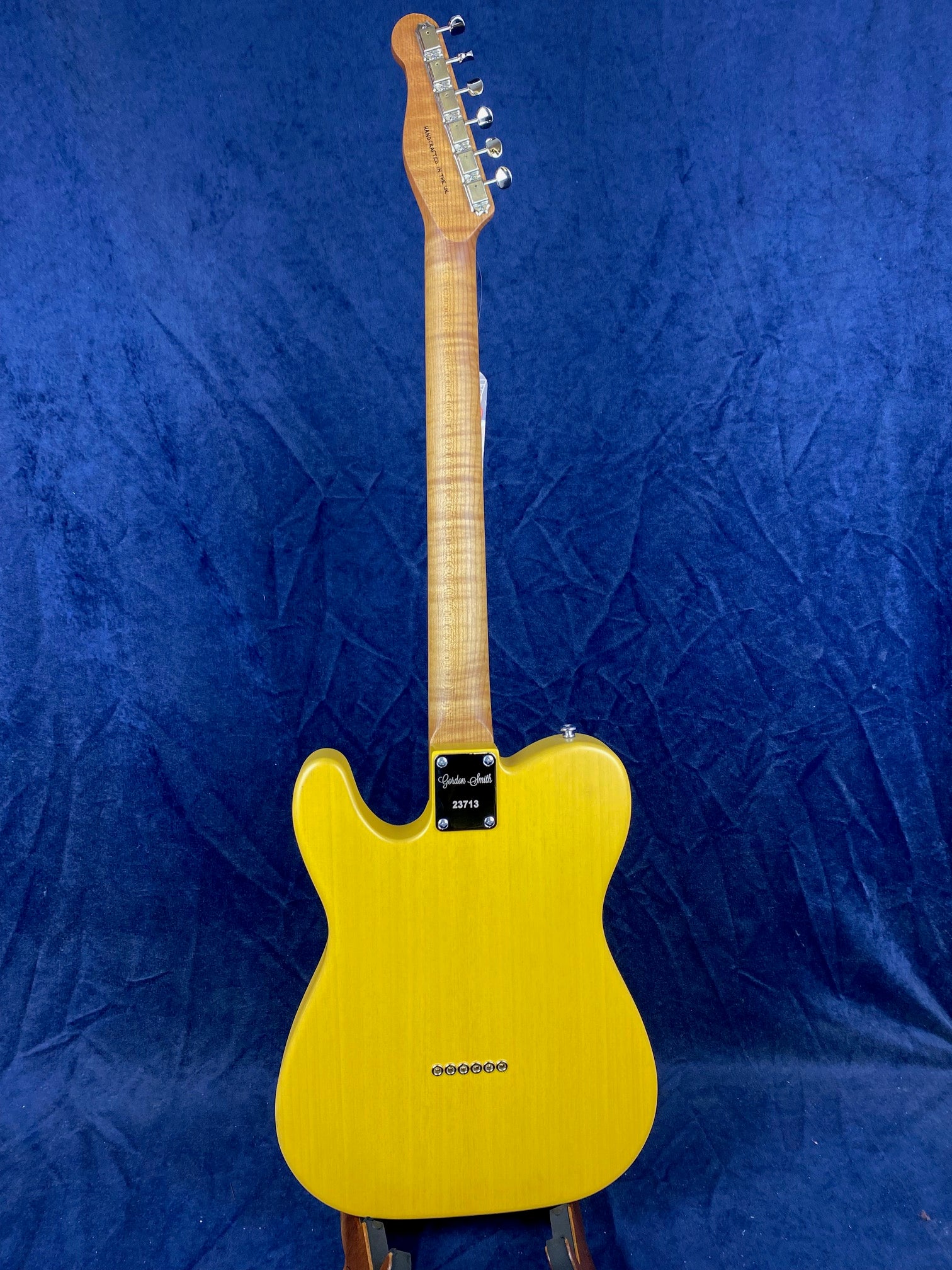 Yellow electric guitar shown from the back, with a smooth contoured body, natural wood neck, and chrome tuning pegs against a dark blue background.
