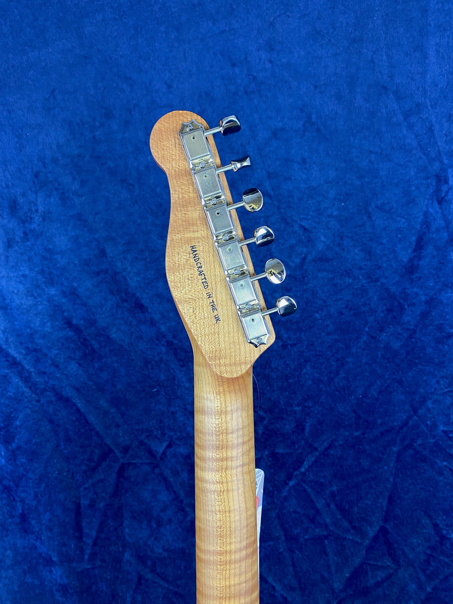Electric guitar neck and headstock seen from the back, with six metal tuners and wavy wood grain, set against a deep blue background.