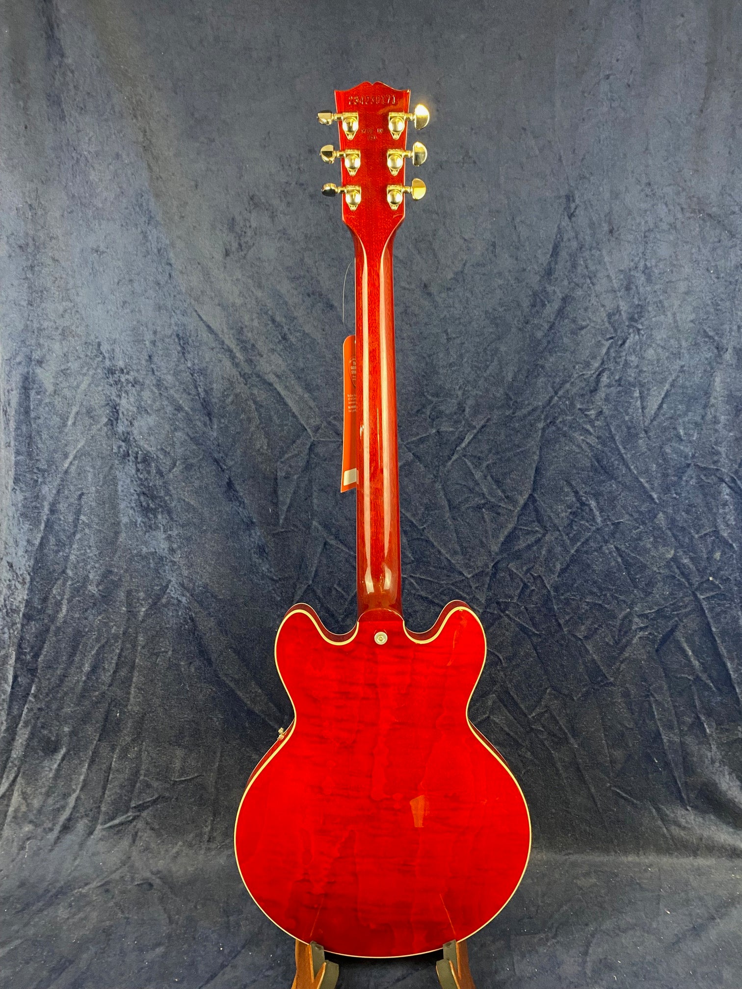 A cherry red semi-hollow electric guitar is shown from the back, highlighting its glossy finish, gold tuning pegs, and double cutaway body shape.