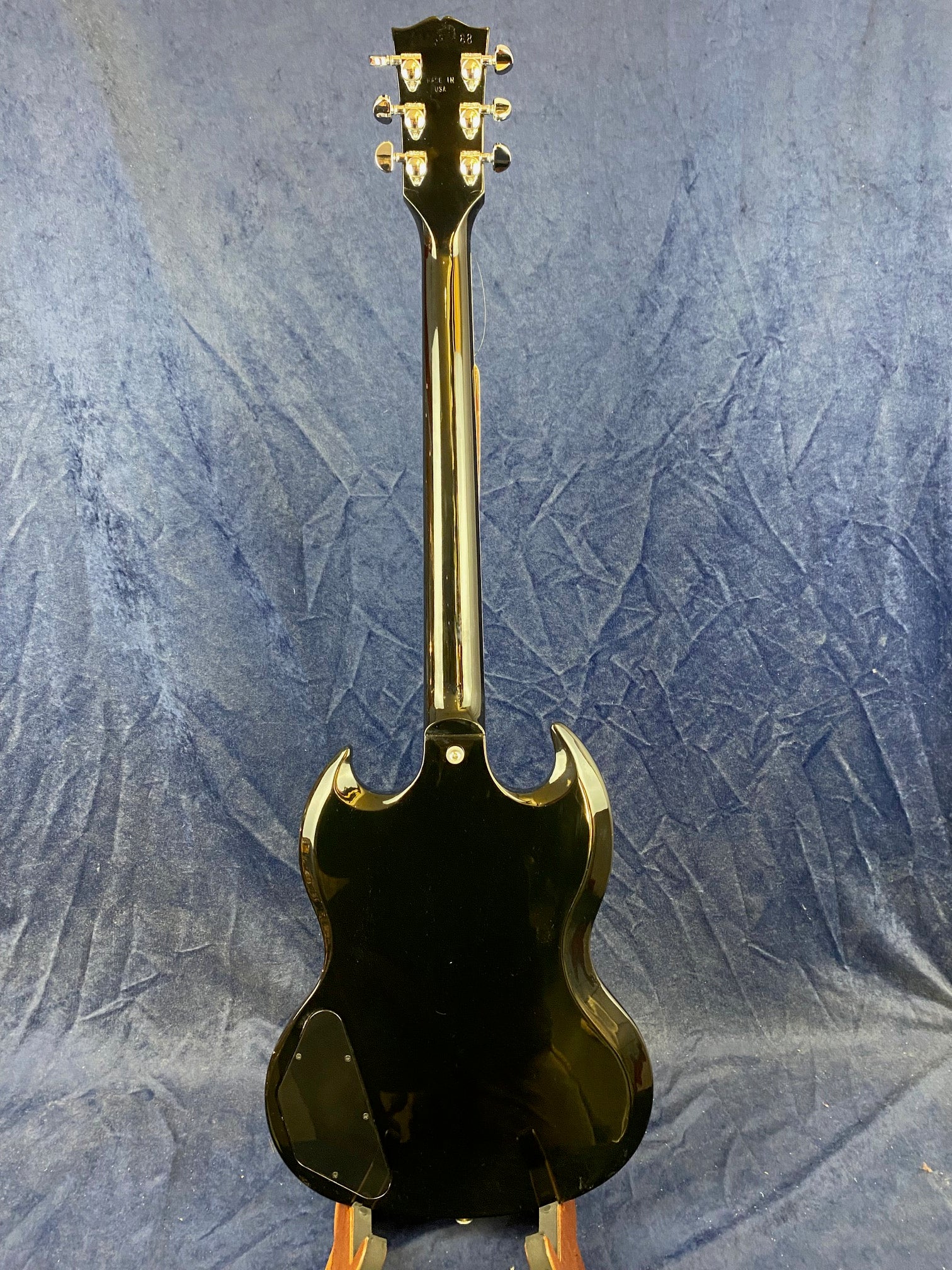 Black Gibson electric guitar shown from the back, with a glossy finish, chrome tuners, and double cutaway body against a blue textured background.