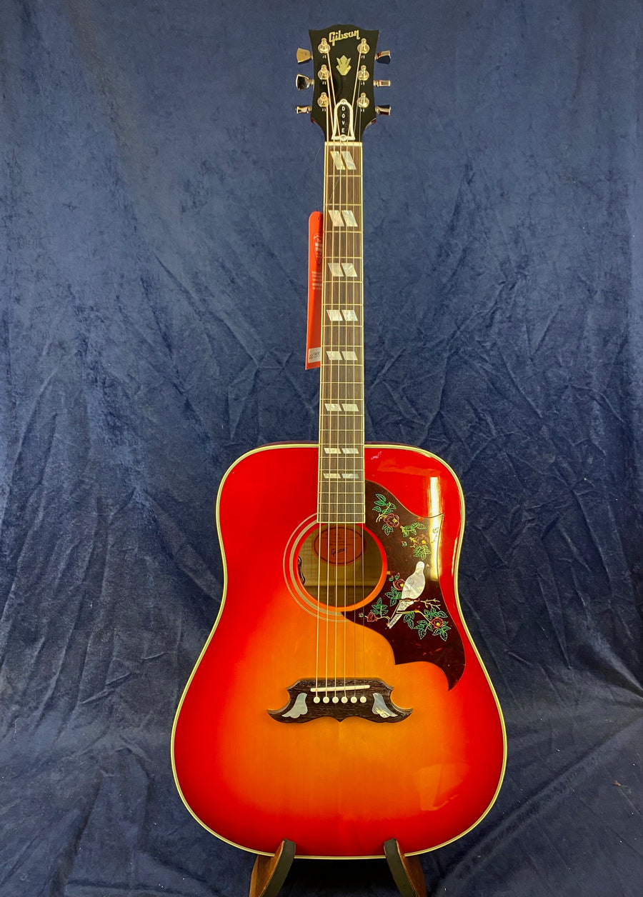 Red-orange Gibson acoustic guitar with a floral pickguard featuring a white dove and green leaves, displayed upright against a dark blue background.