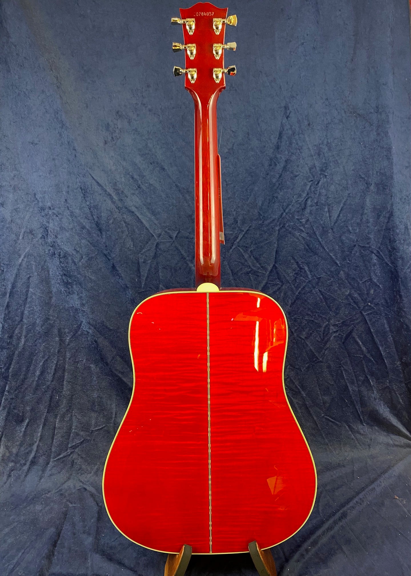 Bright red acoustic guitar shown from the back, with cream binding along the edges and gold tuning pegs, set against a dark blue background.
