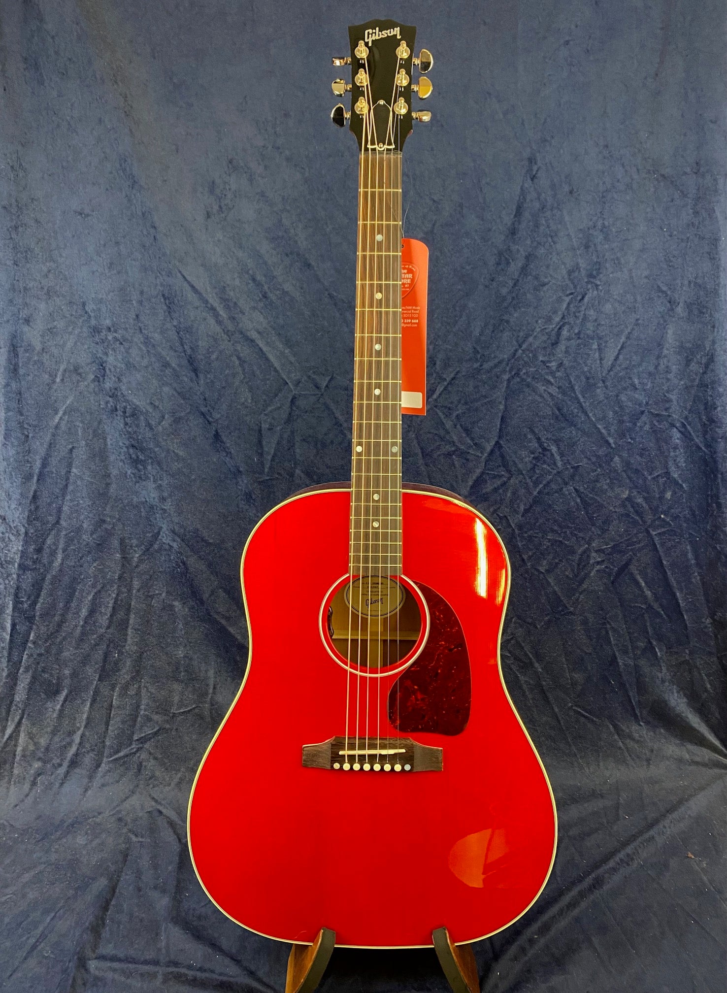 Gibson acoustic guitar with a glossy bright red body, dark pickguard, six strings, and silver tuning pegs, standing upright against a dark background.