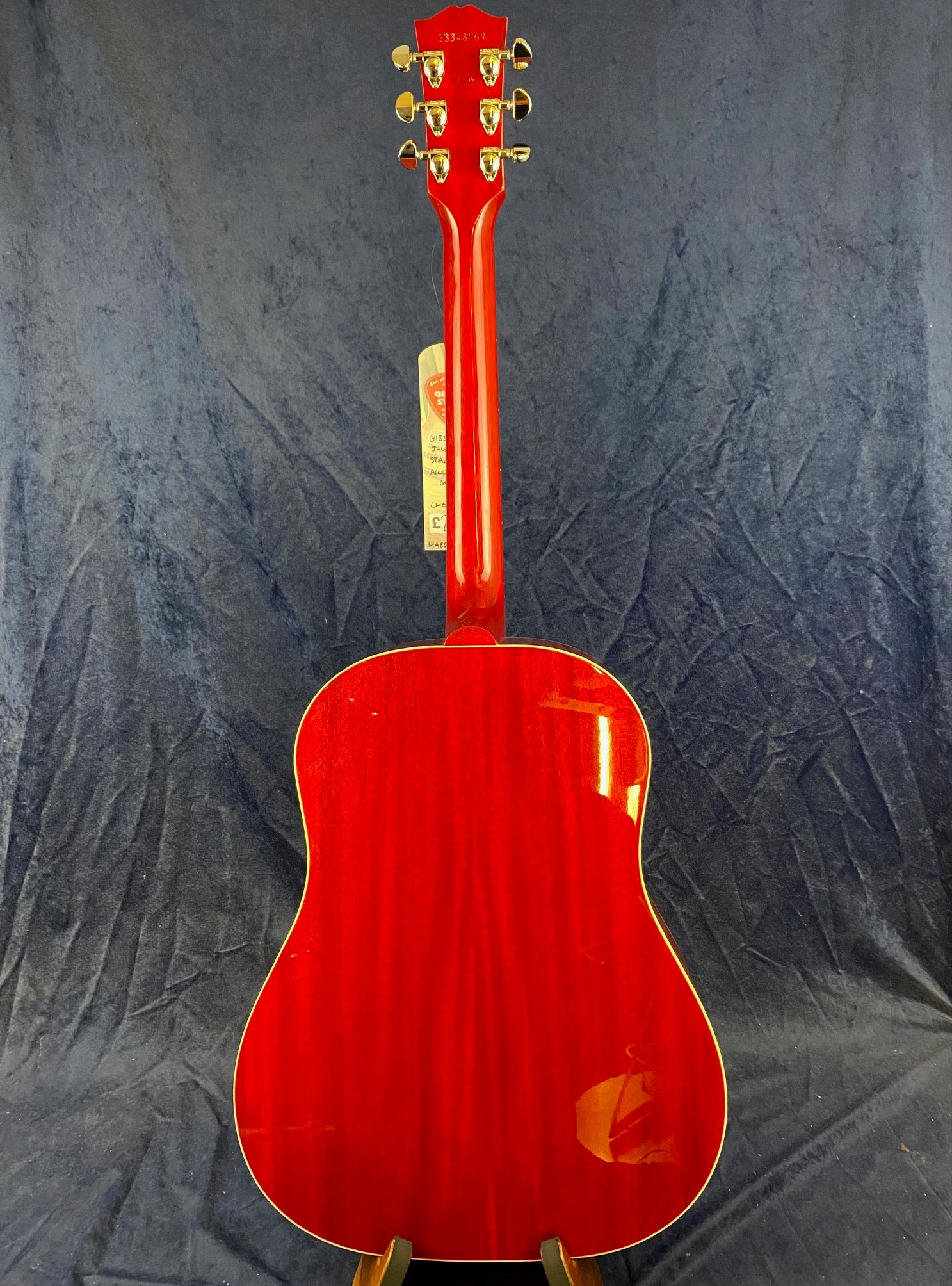 Bright red acoustic guitar shown from the back with gold tuning pegs, glossy finish, and a slightly curved body. A price tag hangs from the headstock.