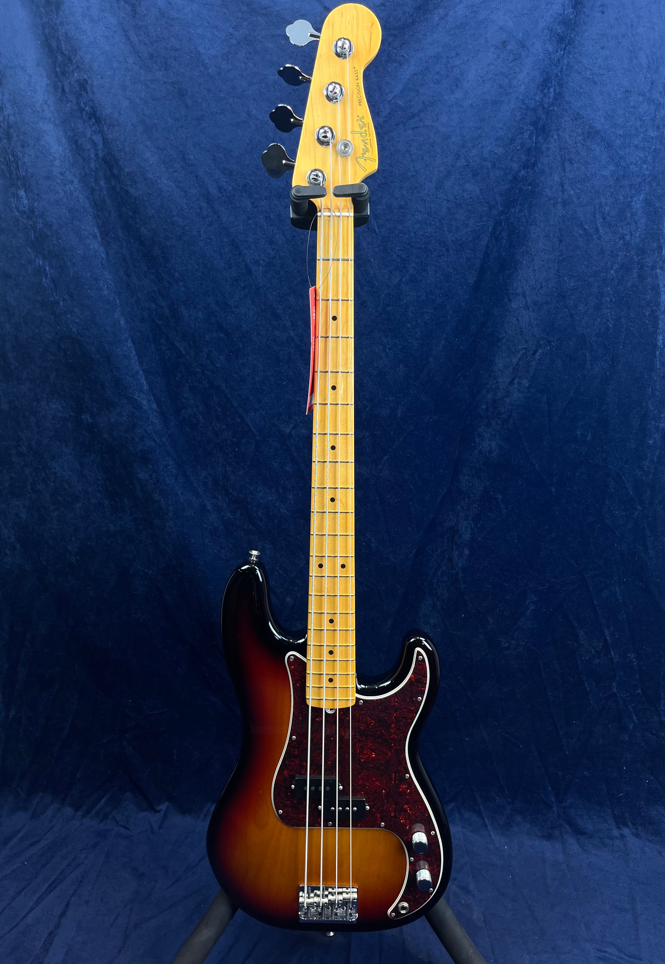 Sunburst finish Fender bass guitar with a tortoiseshell pickguard, four strings, and a maple neck, displayed upright against a dark blue background.