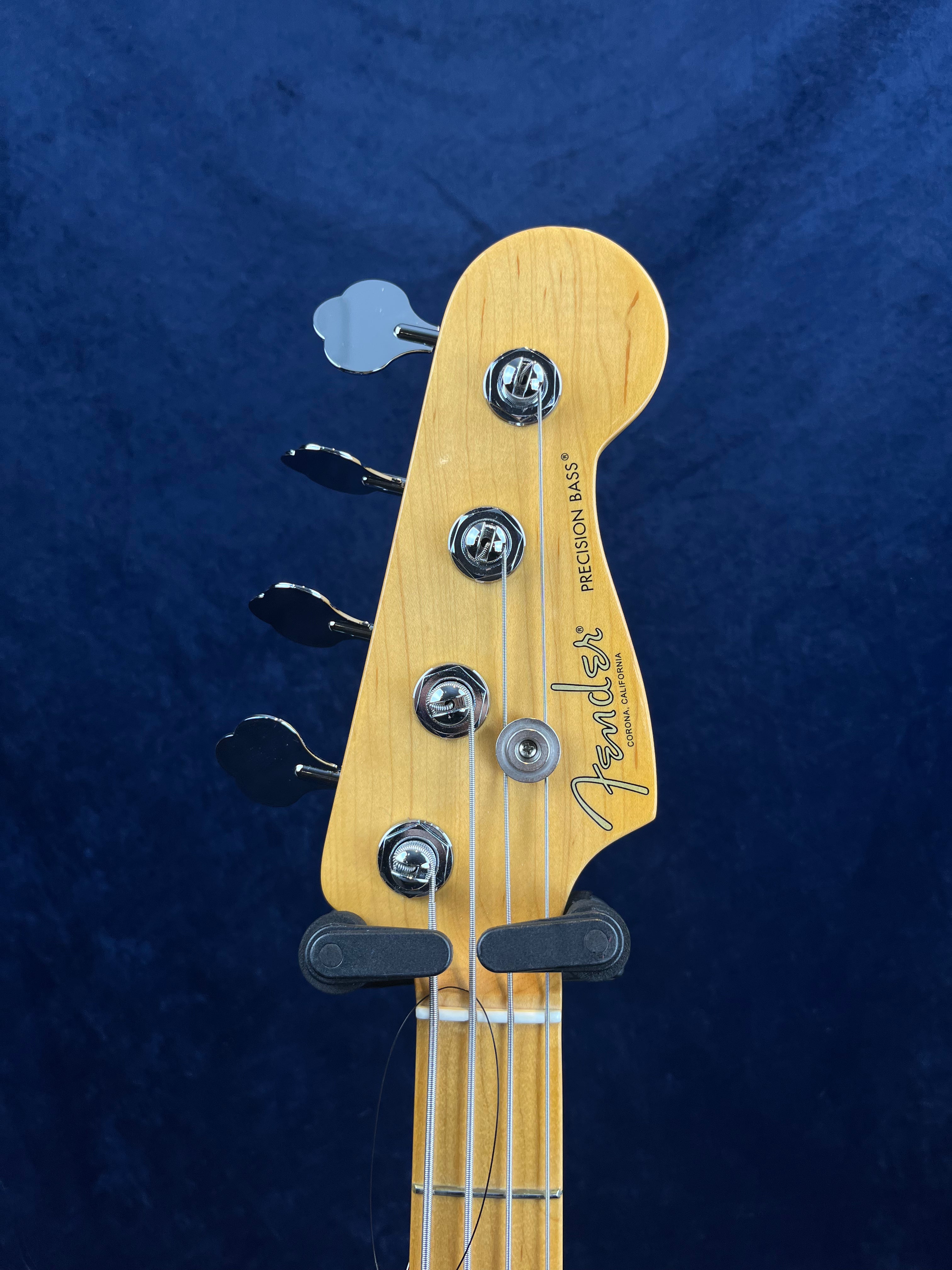 Light wood Fender Precision Bass headstock with four tuning pegs, metal strings, and black logo text against a dark blue background.