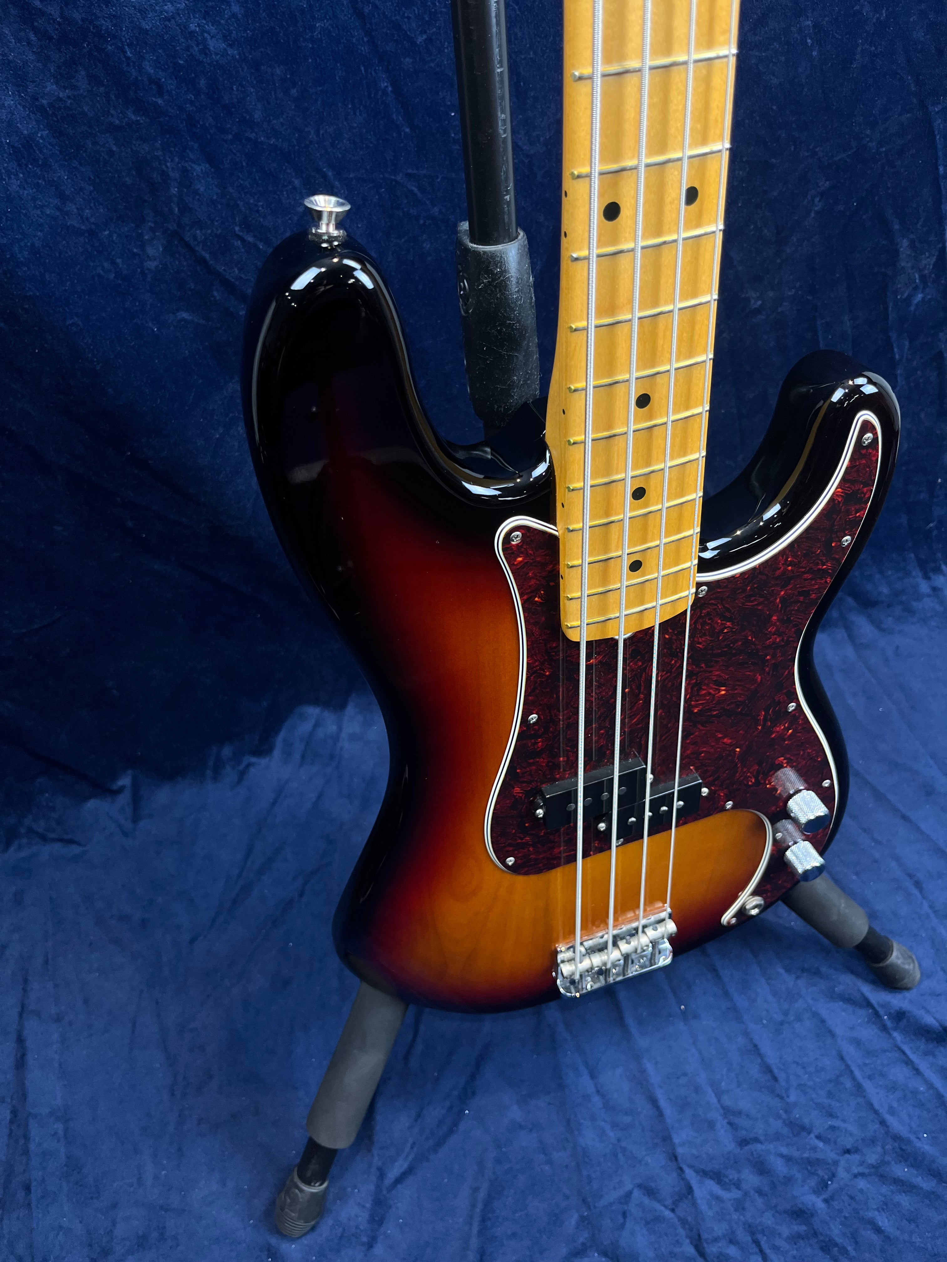 Four-string sunburst bass guitar with a tortoiseshell pickguard, maple neck, and chrome hardware, resting on a black stand against a dark blue background.