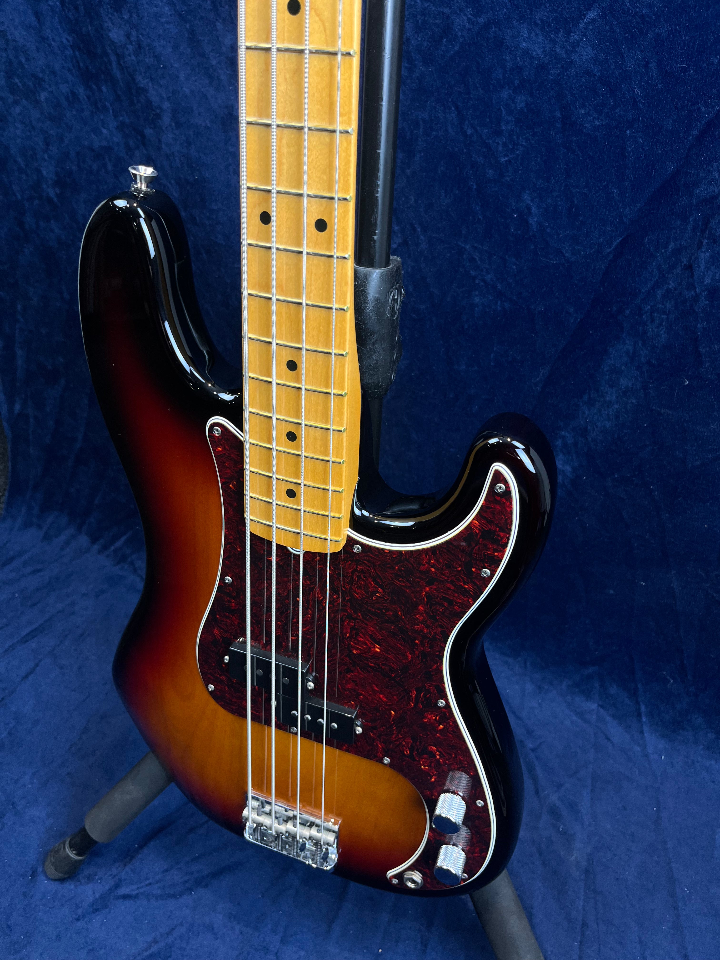 Sunburst finish bass guitar with a tortoiseshell pickguard, maple neck, four strings, and chrome control knobs, displayed on a stand against blue fabric.