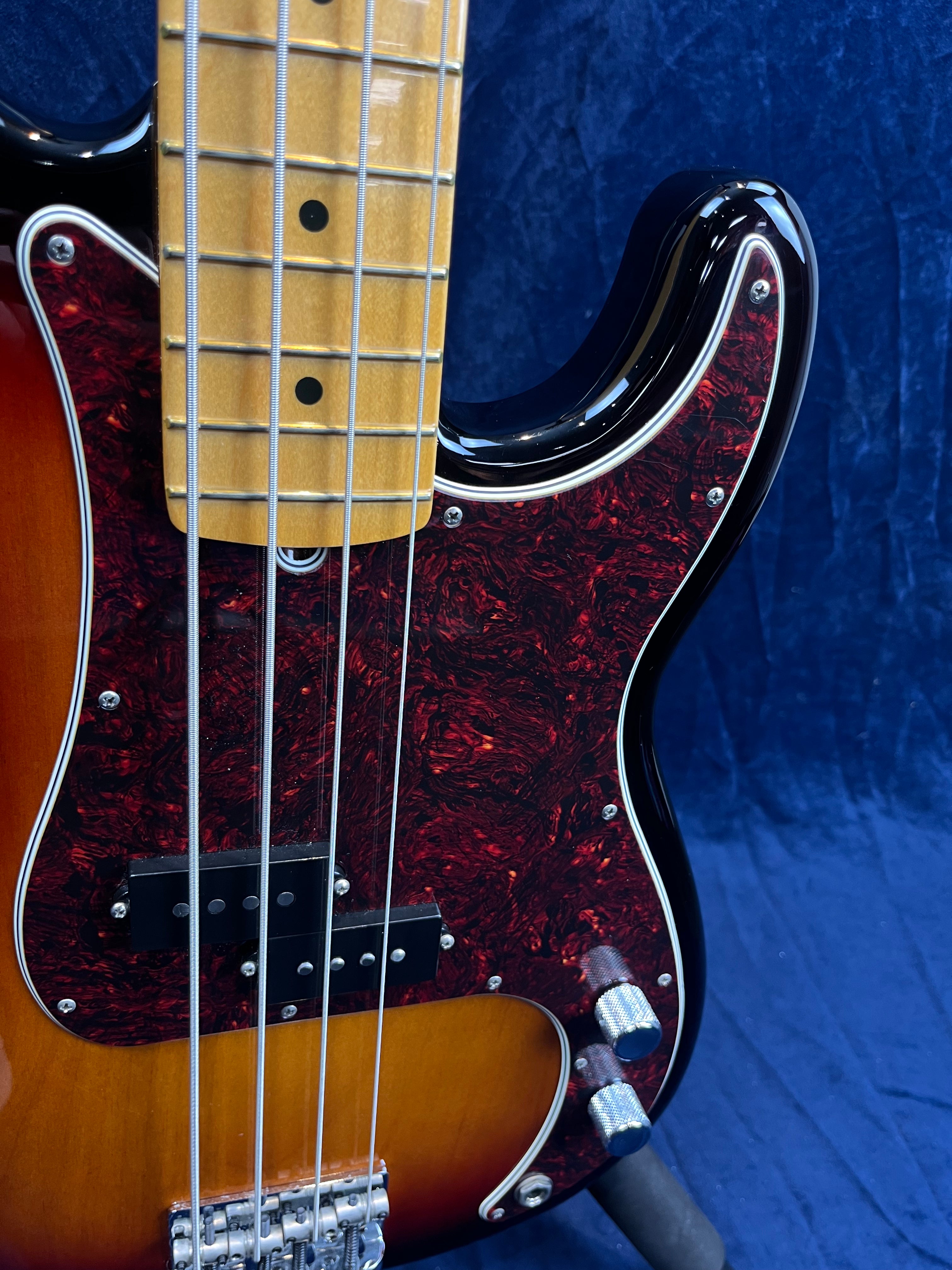 Sunburst bass guitar with a maple neck, black and red tortoiseshell pickguard, chrome control knobs, and four strings against a blue background.