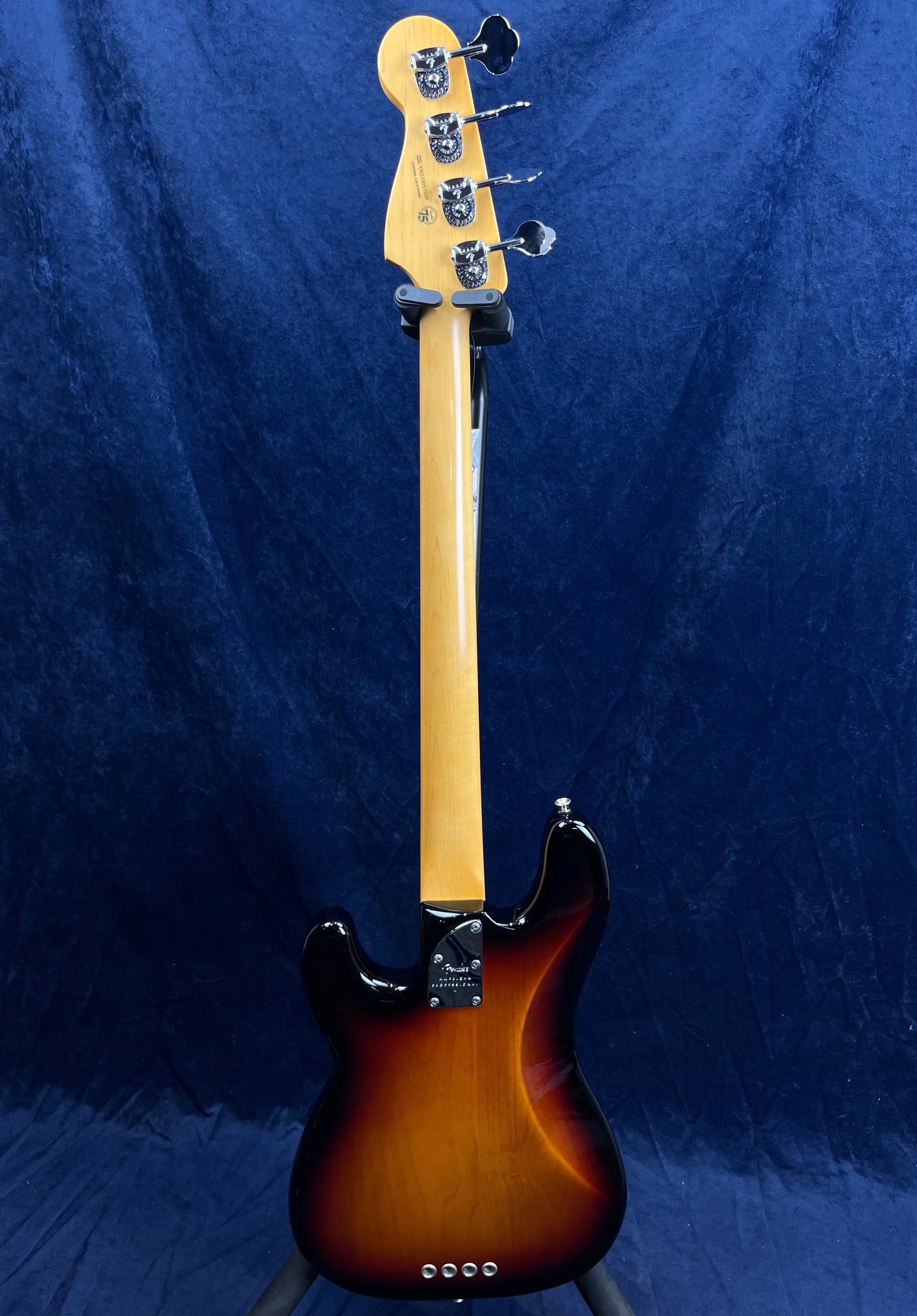 Sunburst-finish bass guitar shown from the back, with a maple neck and four open-gear tuners, resting upright against a dark blue background.