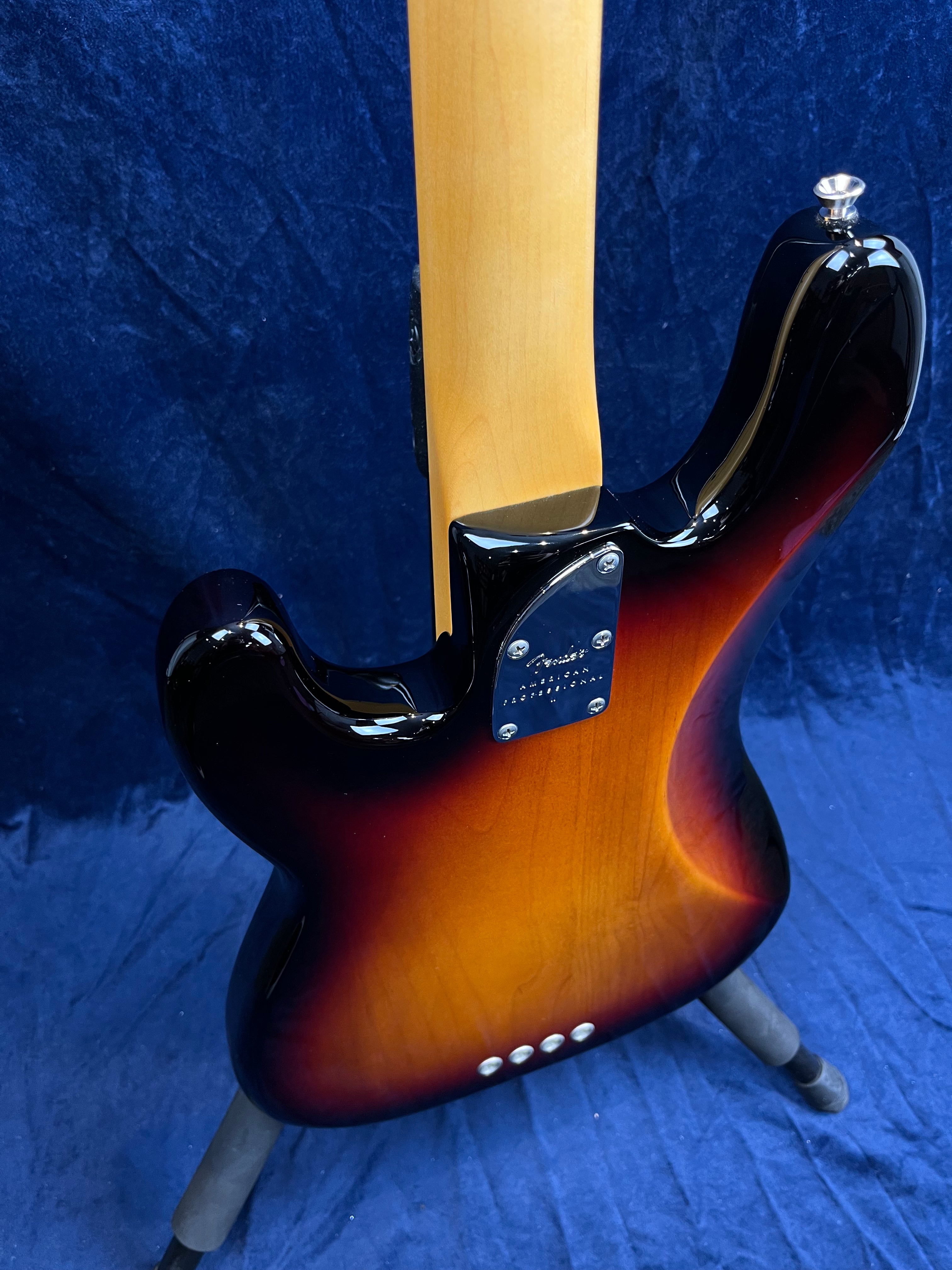 Sunburst finish bass guitar viewed from the back, showing a maple neck, chrome neck plate, and four string ferrules on a dark blue background.