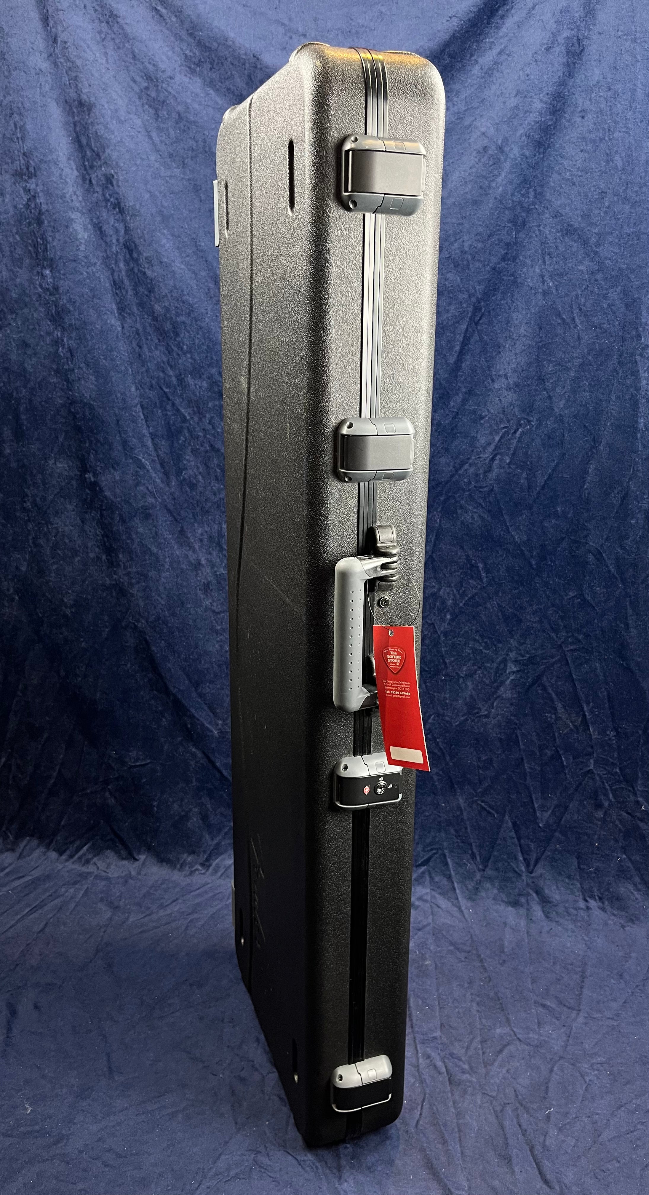 Tall, rectangular hard-shell bass guitar case stands upright with four latches and a gray handle; a red tag hangs from the handle.