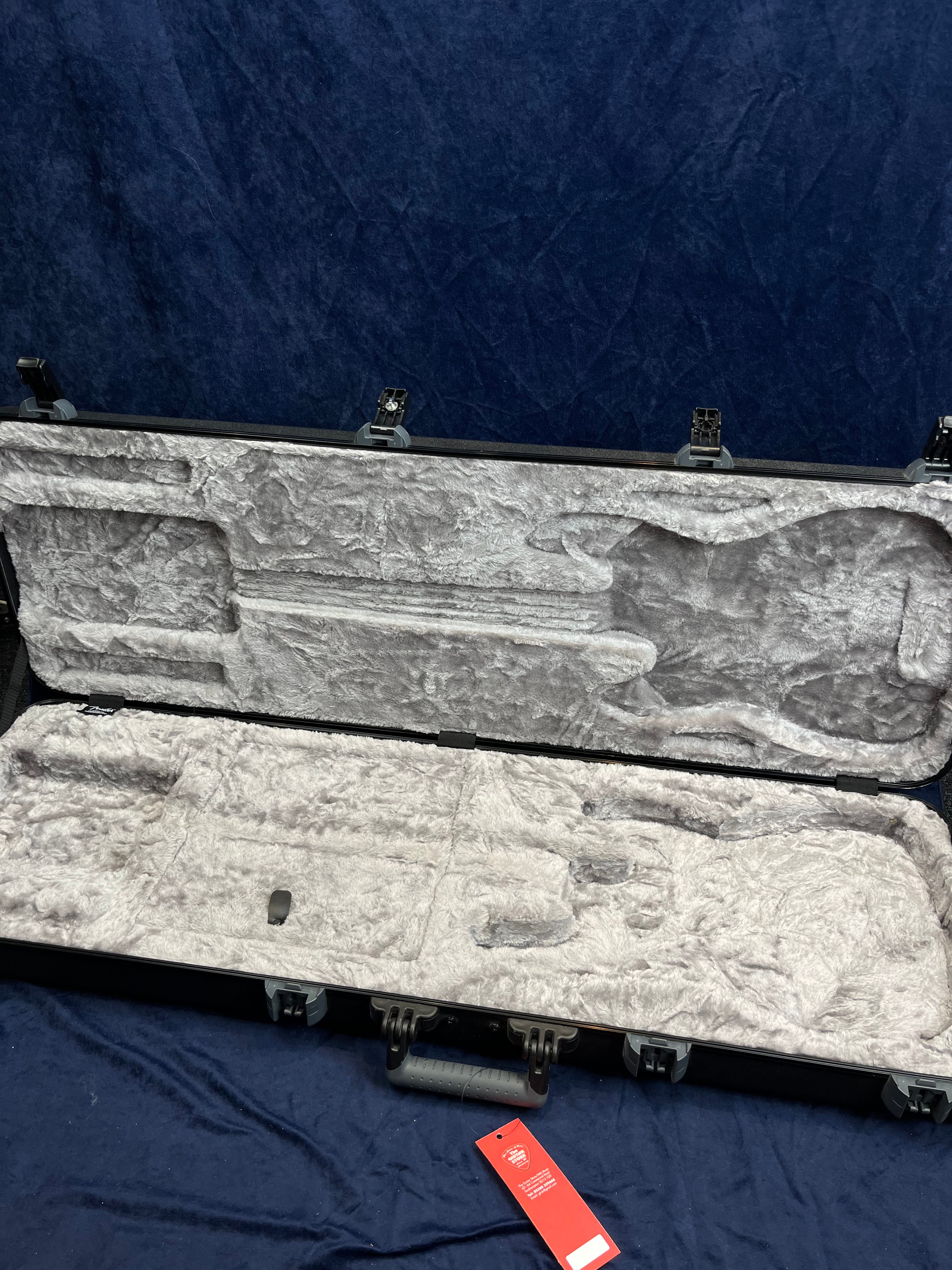 Open hard shell bass guitar case lined with gray plush fabric, showing molded spaces for the instrument and accessories, empty and lying on a dark cloth.