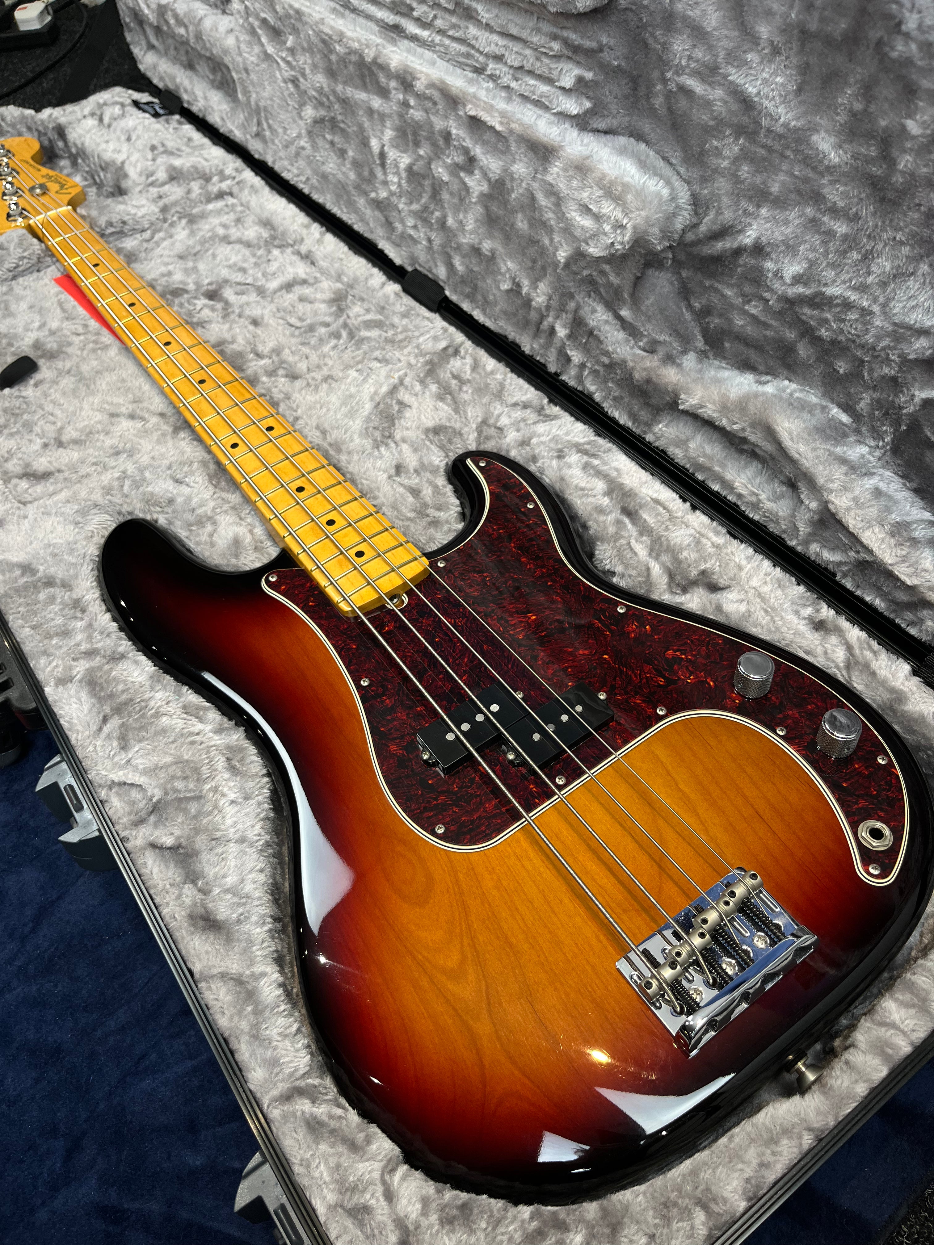 Sunburst-finish bass guitar with a tortoiseshell pickguard, maple neck, and chrome hardware resting in a gray plush-lined hard case.