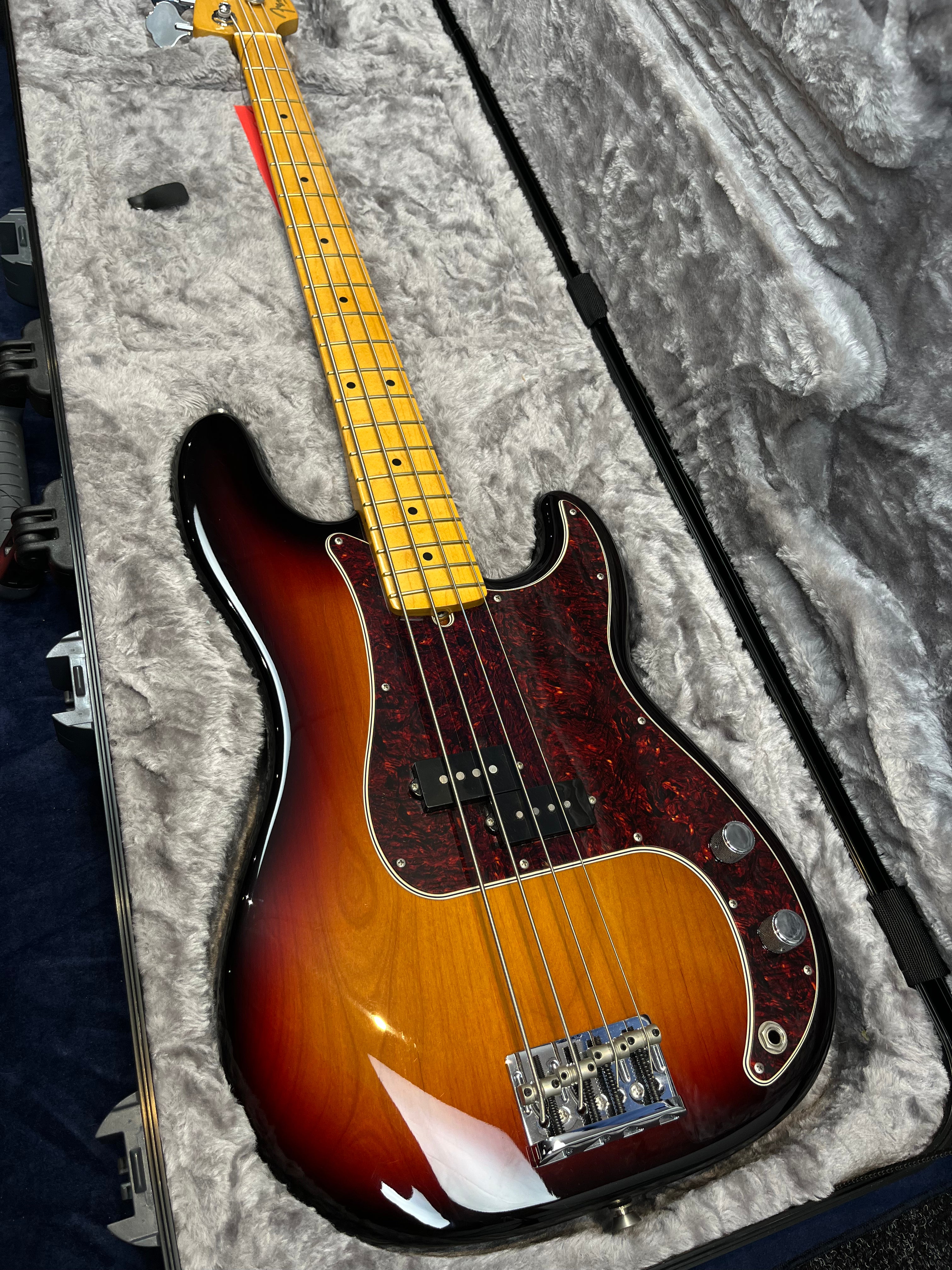 Sunburst electric bass guitar with a maple neck and dark red tortoiseshell pickguard, resting in a gray plush-lined hard case.