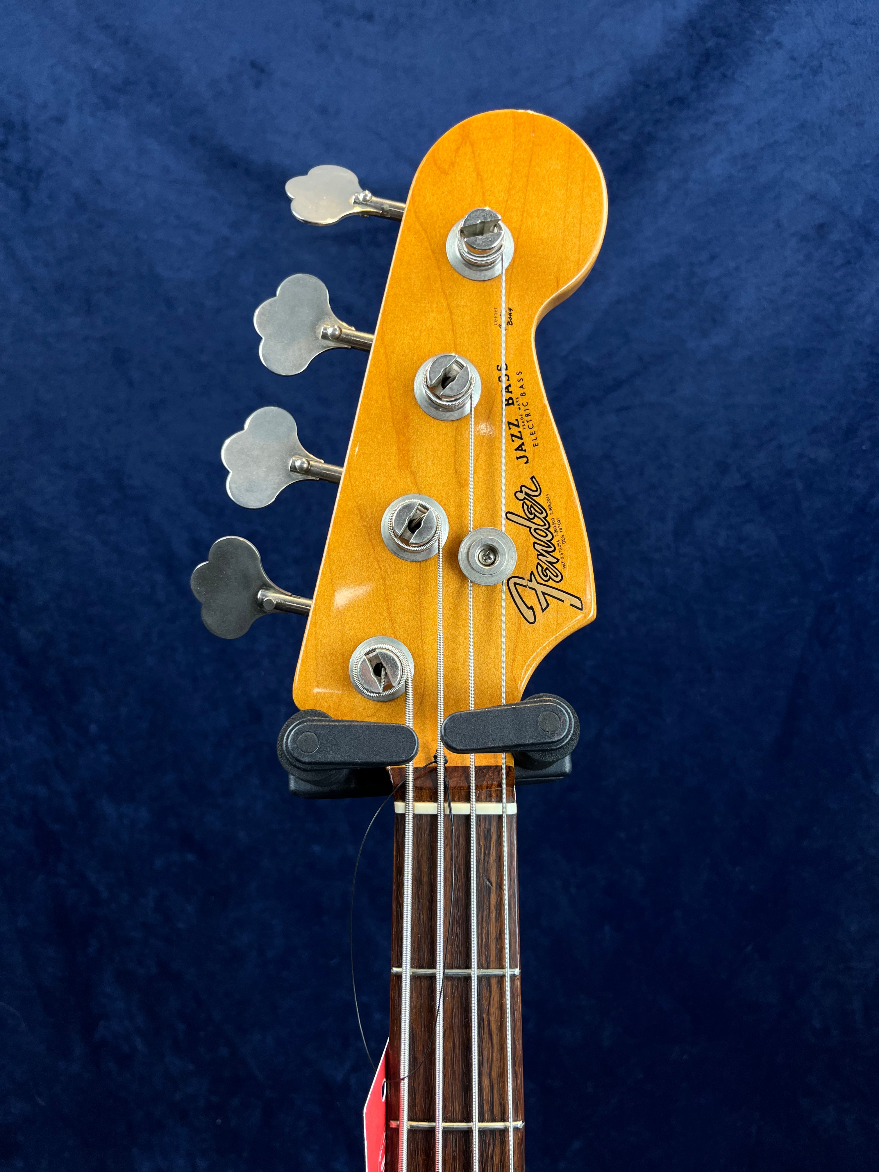 Fender Jazz Bass guitar headstock with four tuning pegs, visible strings, and part of the rosewood fretboard against a blue background.