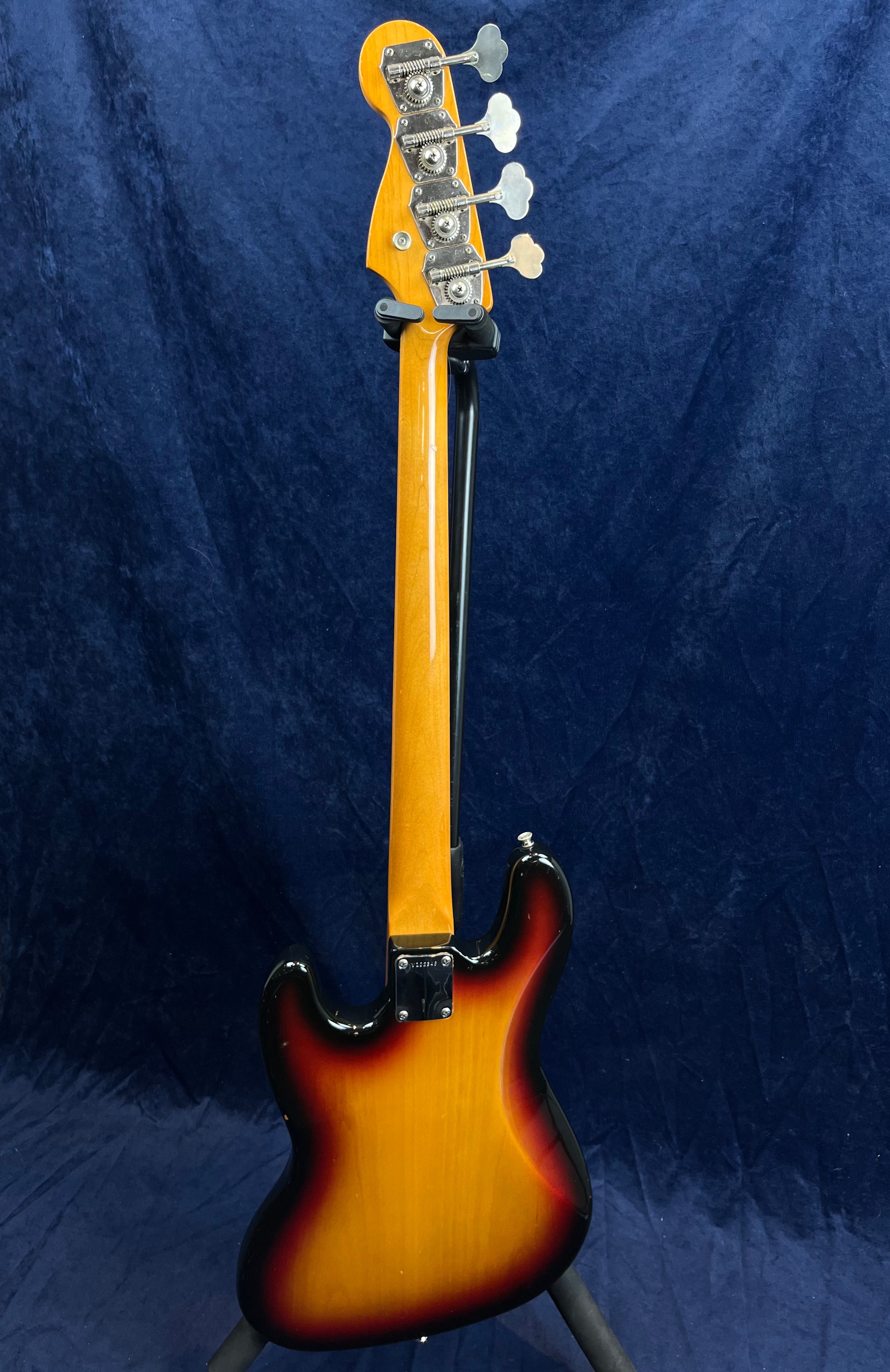 Sunburst finish bass guitar viewed from the back, showing the neck, tuners, and curved body against a dark blue background.