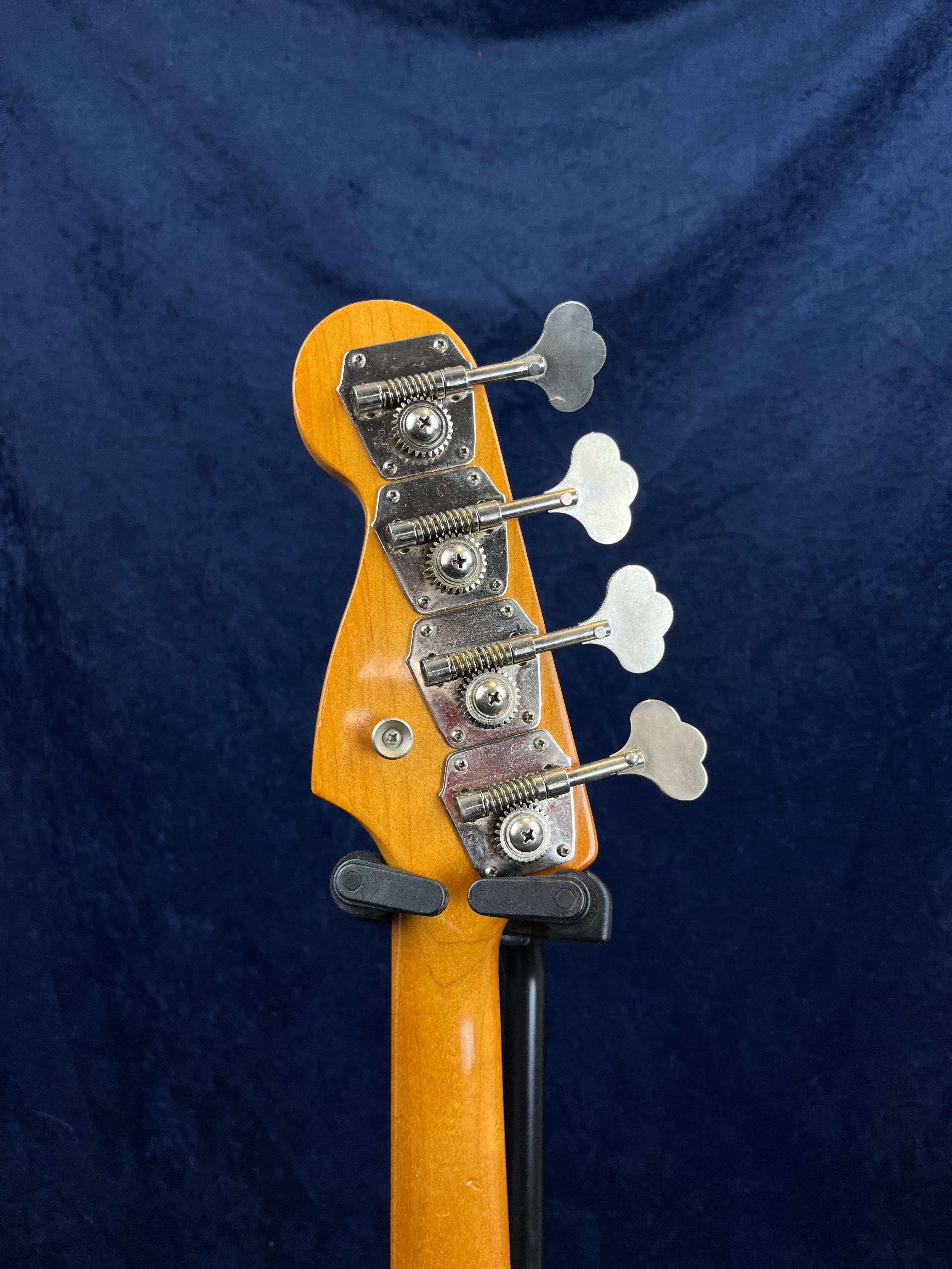 Bass guitar headstock showing four metal tuning pegs on a light wood finish, set against a dark blue background.