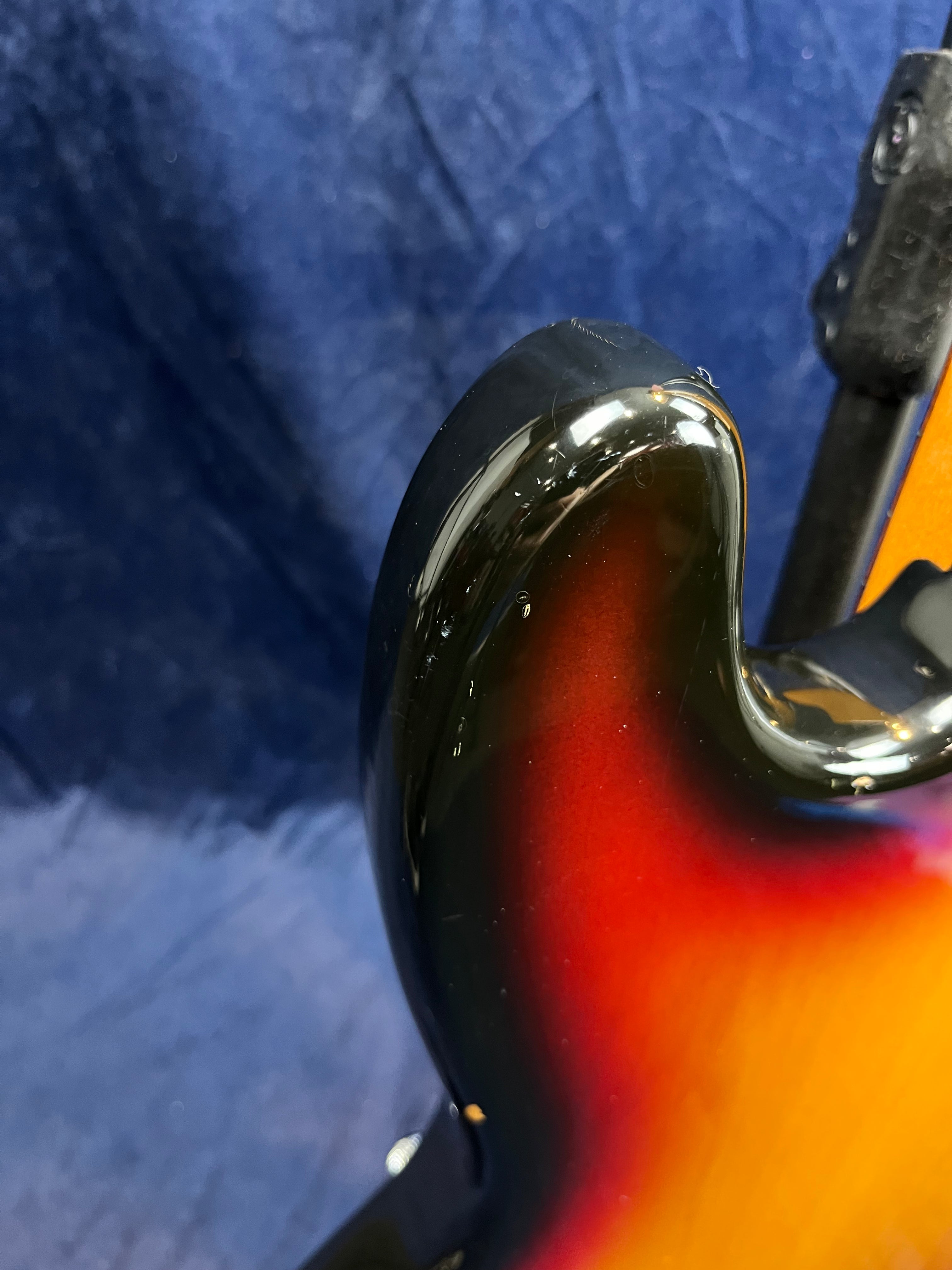 A close-up of a bass guitar body shows a glossy sunburst finish with yellow, red, and black colors, and minor chips near the edge.