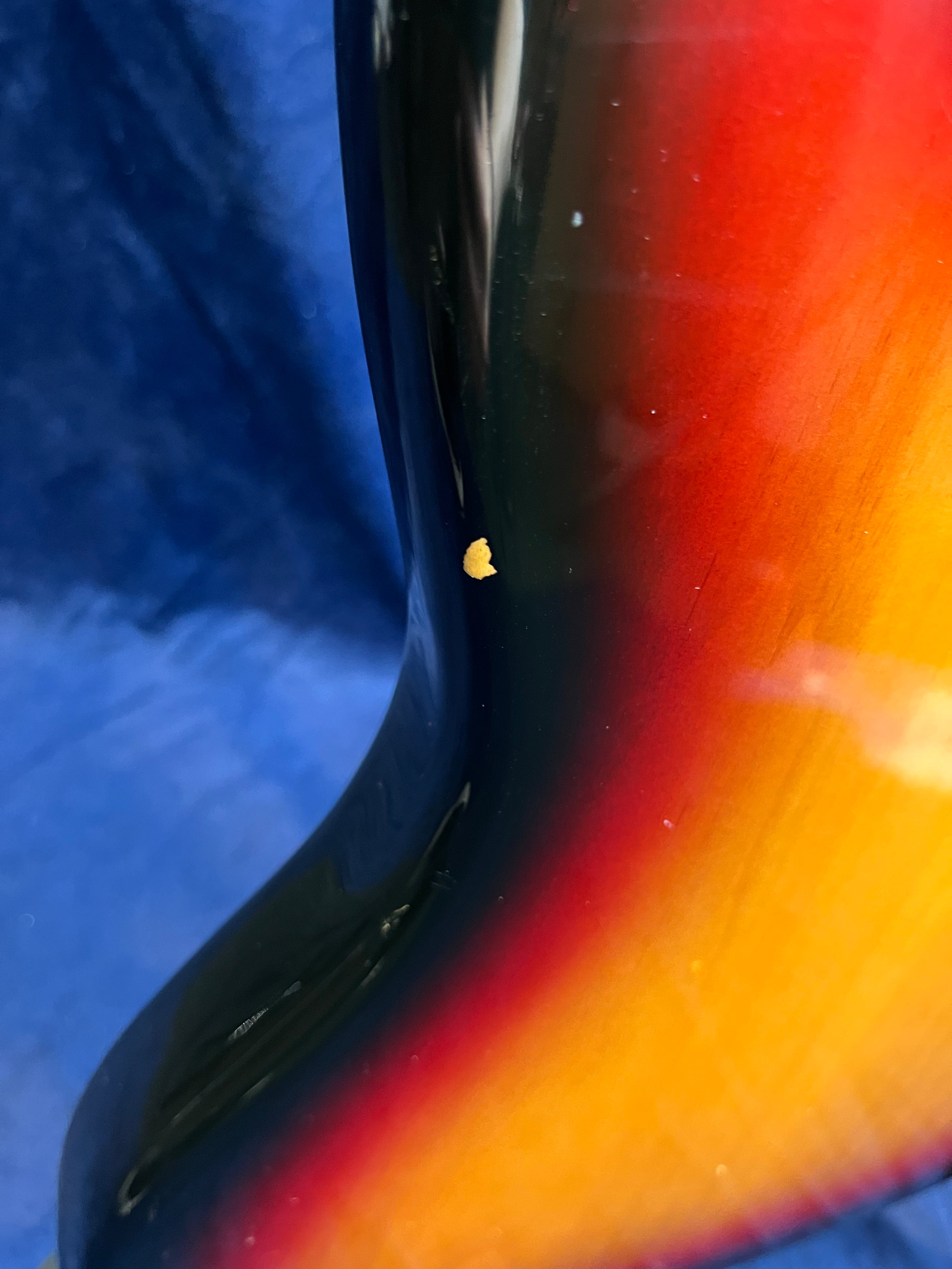Sunburst finish bass guitar body with a small chip in the paint exposing wood, set against a blue background.