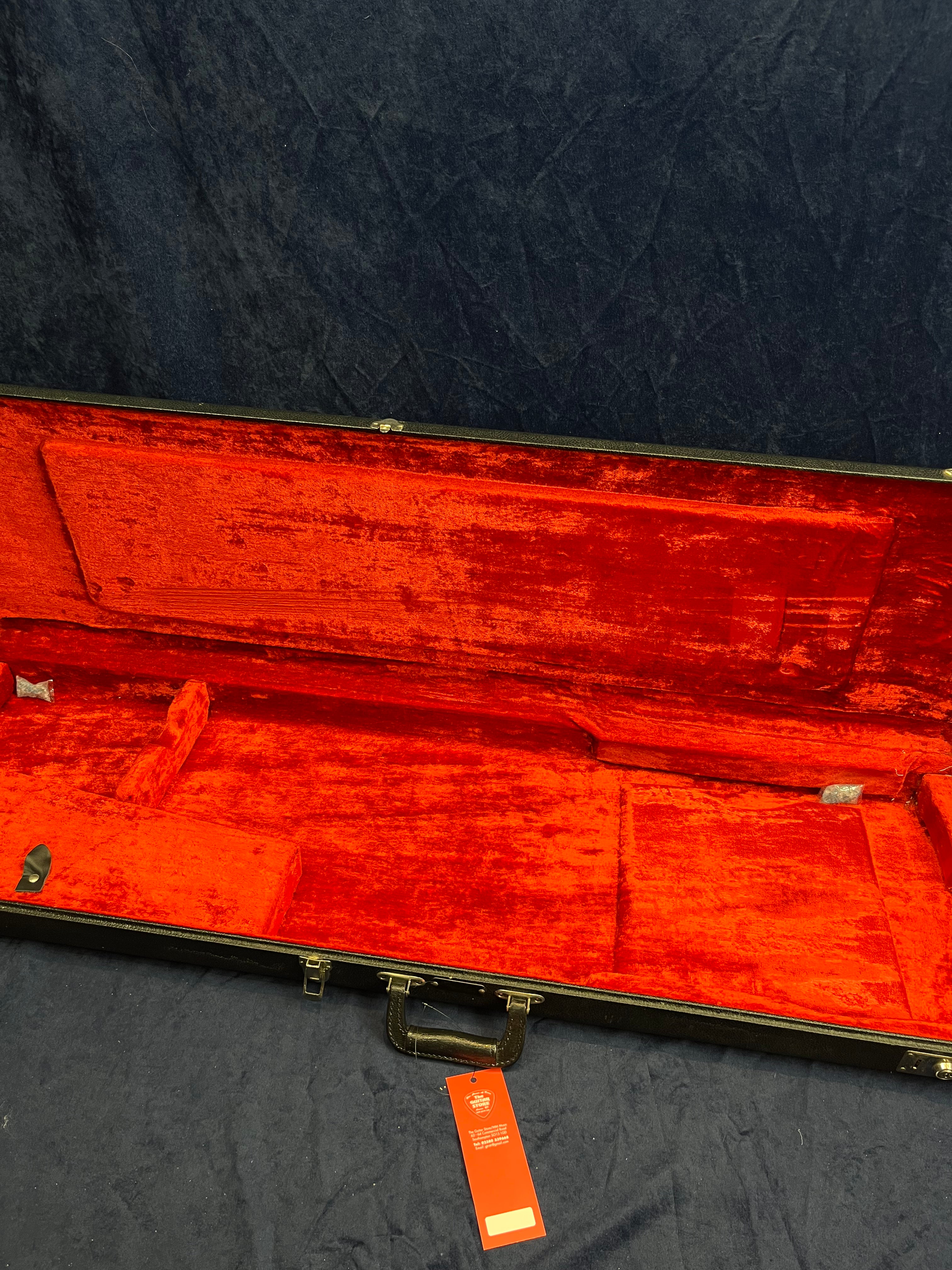 Empty rectangular bass guitar case with bright red plush interior, black outer edges, handle, and a red tag attached to the latch area.