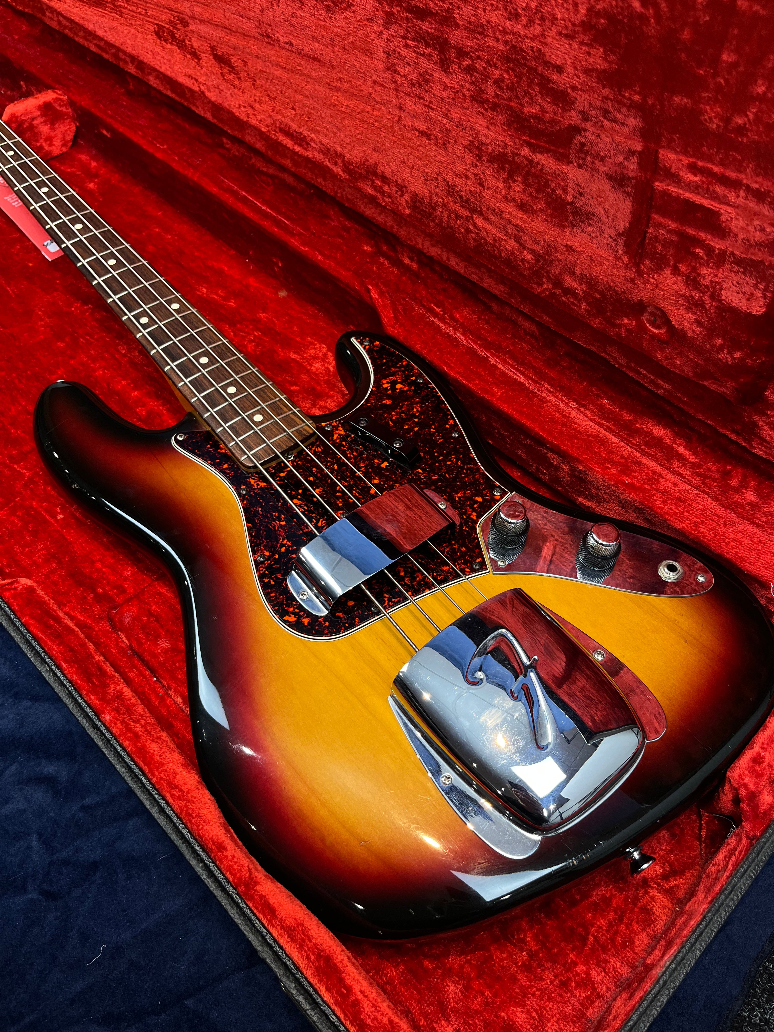 Sunburst Fender bass guitar with chrome pickup and bridge covers, tortoiseshell pickguard, and two control knobs, resting in a red velvet case.